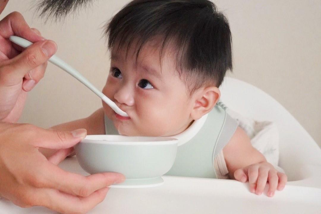 A baby in a bib sitting in a high chair, being fed with a spoon from a bowl by an adult hand.