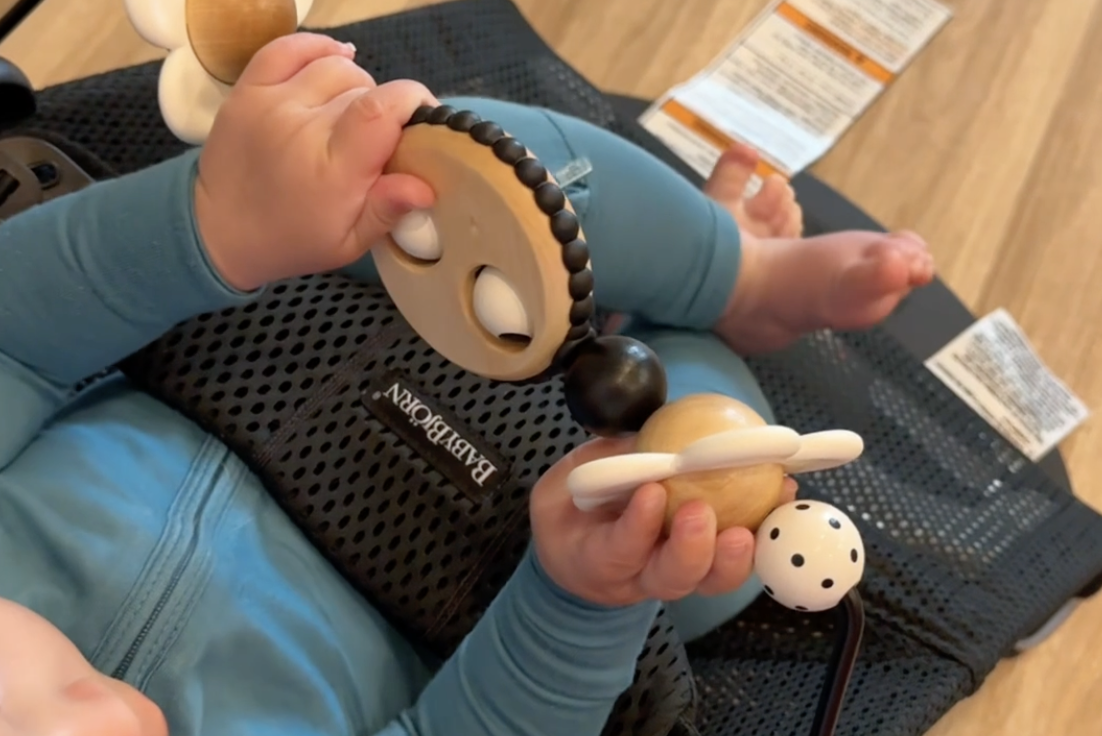 A baby wearing a blue outfit holds a wooden toy with black and white details while sitting in a mesh chair on a wooden floor.