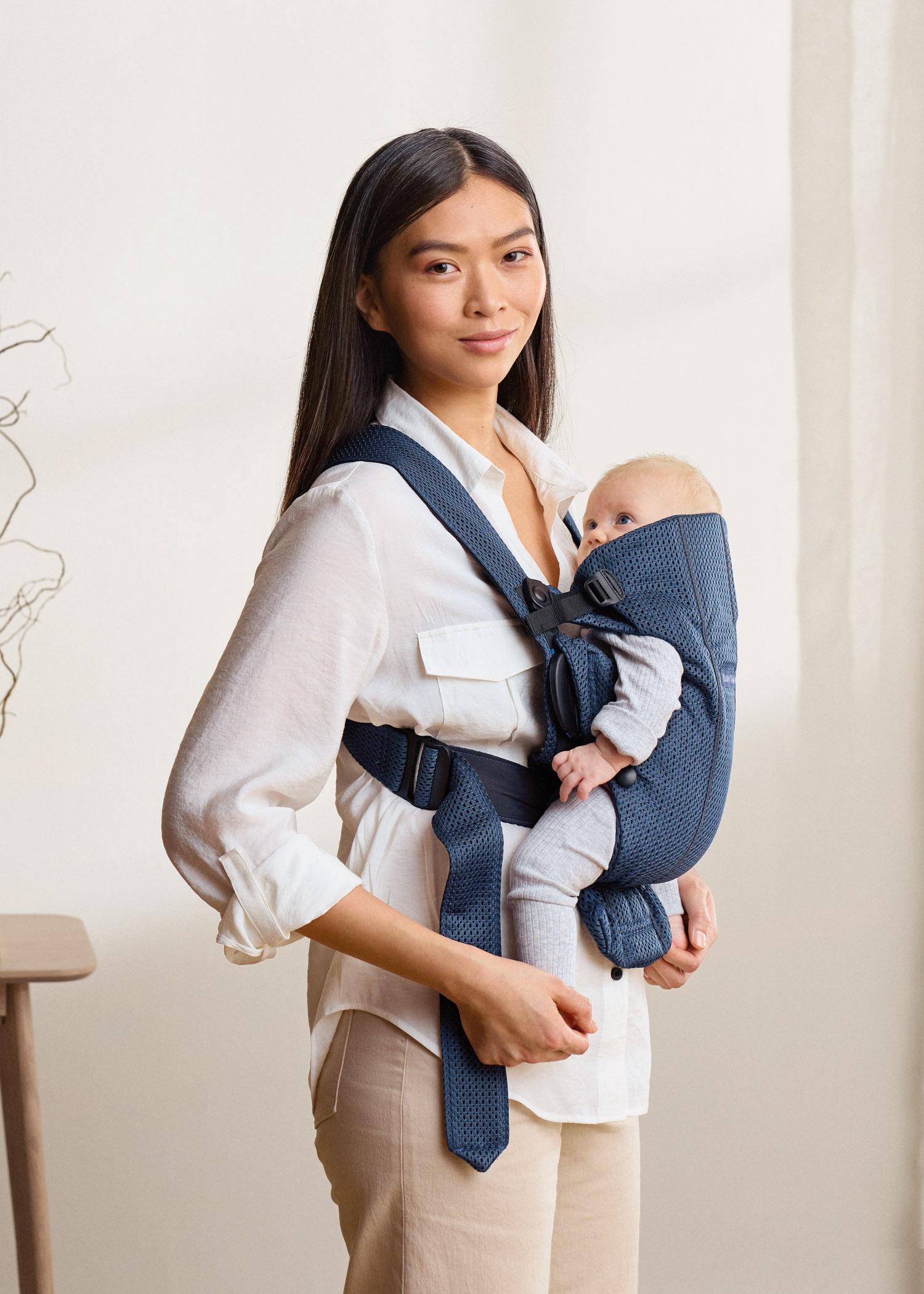 Woman wearing a white shirt carries a baby in a blue carrier. The setting is a softly lit room with light-colored walls.