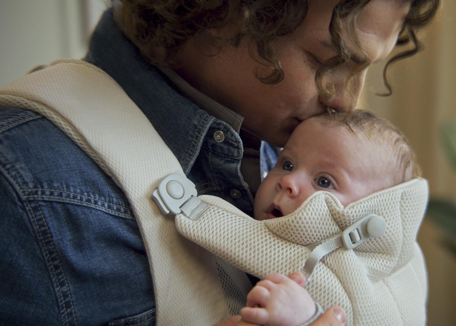 A person wearing a denim shirt kisses the forehead of a baby in a white carrier, who looks upward with wide eyes.