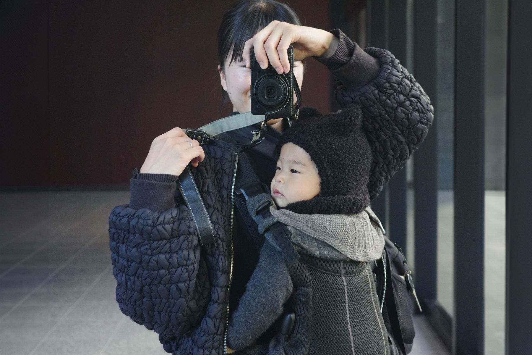 Person taking a mirror selfie while carrying a baby in a front-facing carrier. Both are wearing warm, dark clothing.