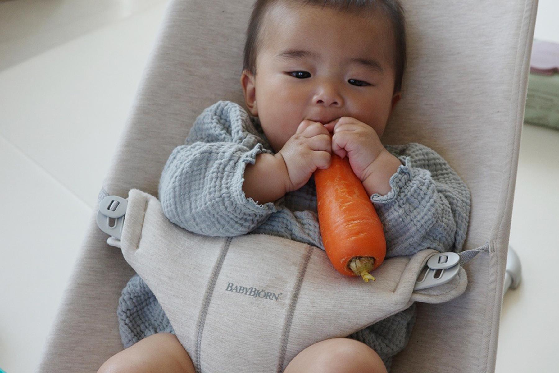 Baby in a gray outfit sits in a beige baby chair, holding and chewing on a large carrot.