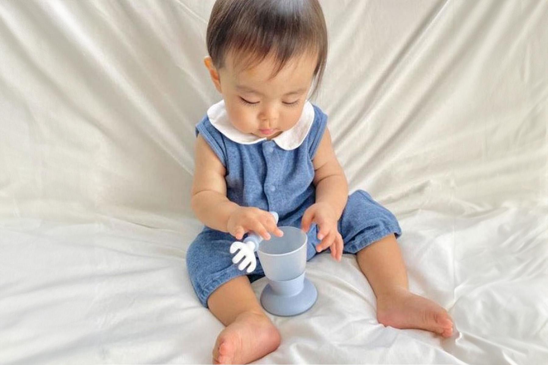 A baby in a blue outfit sits on a white sheet, playing with a light blue cup and a small white object in its hands.
