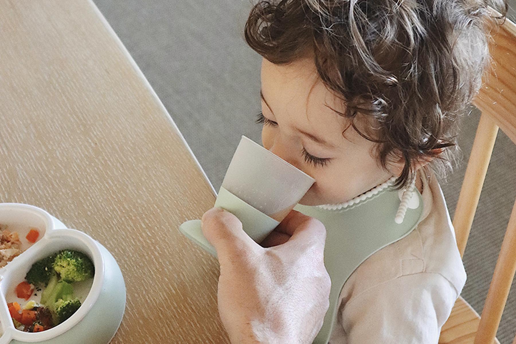 Child with curly hair drinking from a cup held by an adult, sitting at a table with a plate of food including broccoli.