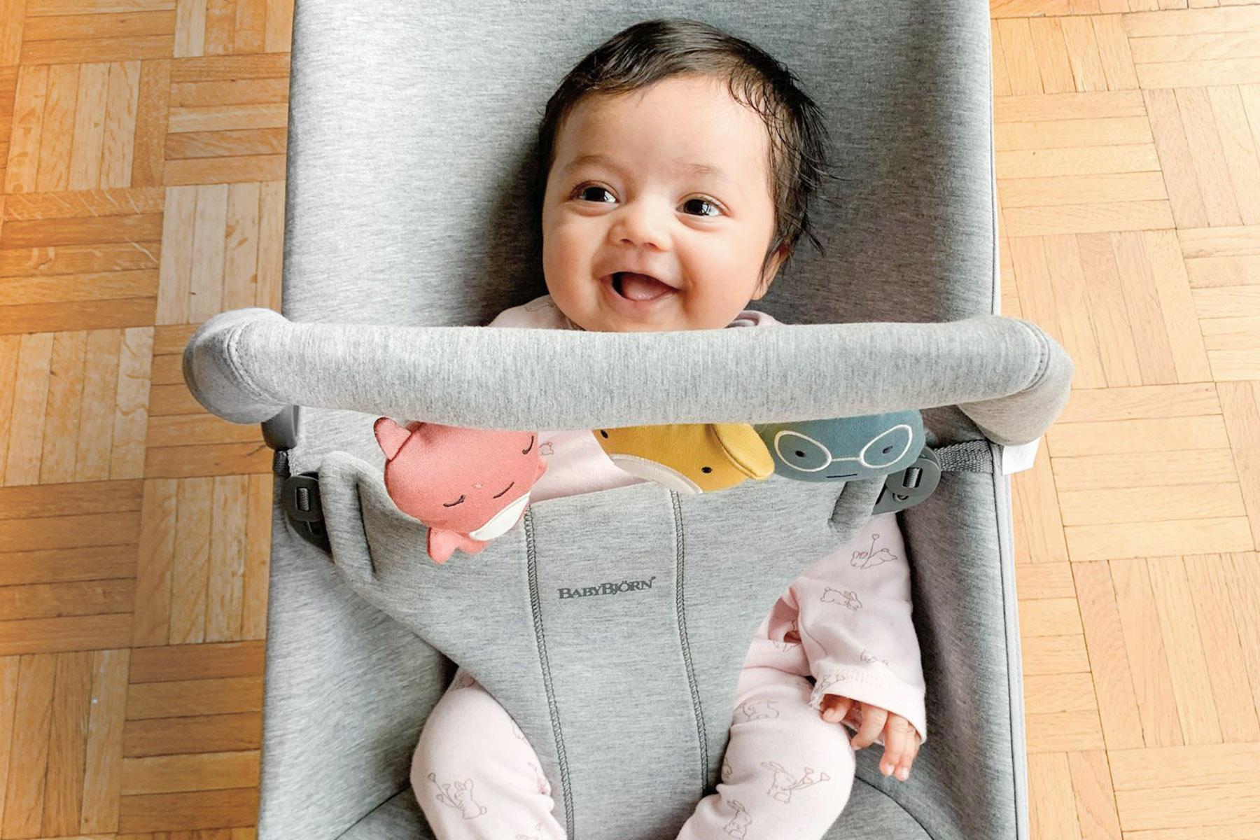 Smiling baby in a gray bouncer wearing pink pajamas, toy bar with plush animals, on a wood parquet floor.