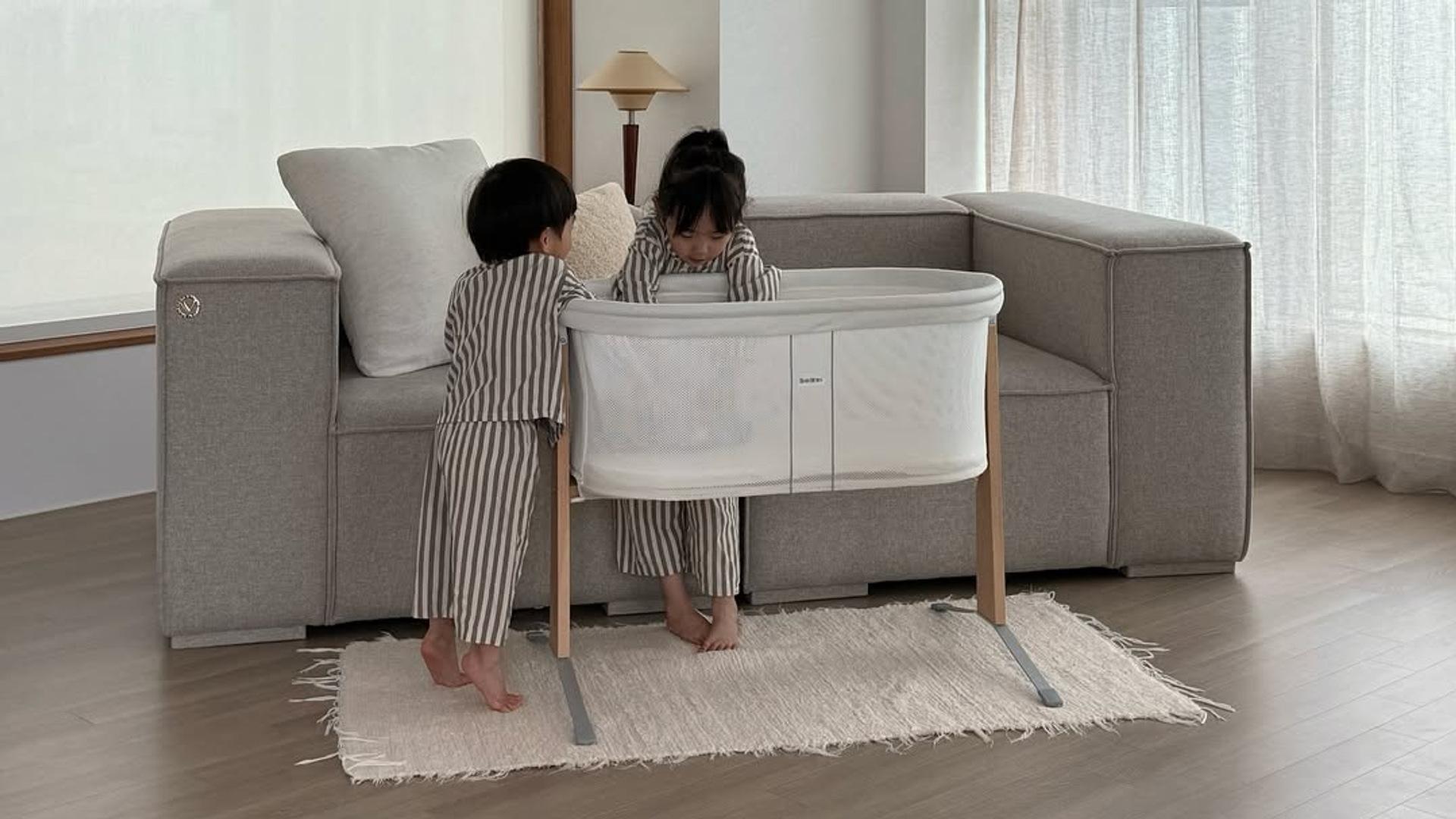 Two children in striped pajamas stand by a white baby cradle in a bright living room with a gray couch and a rug.