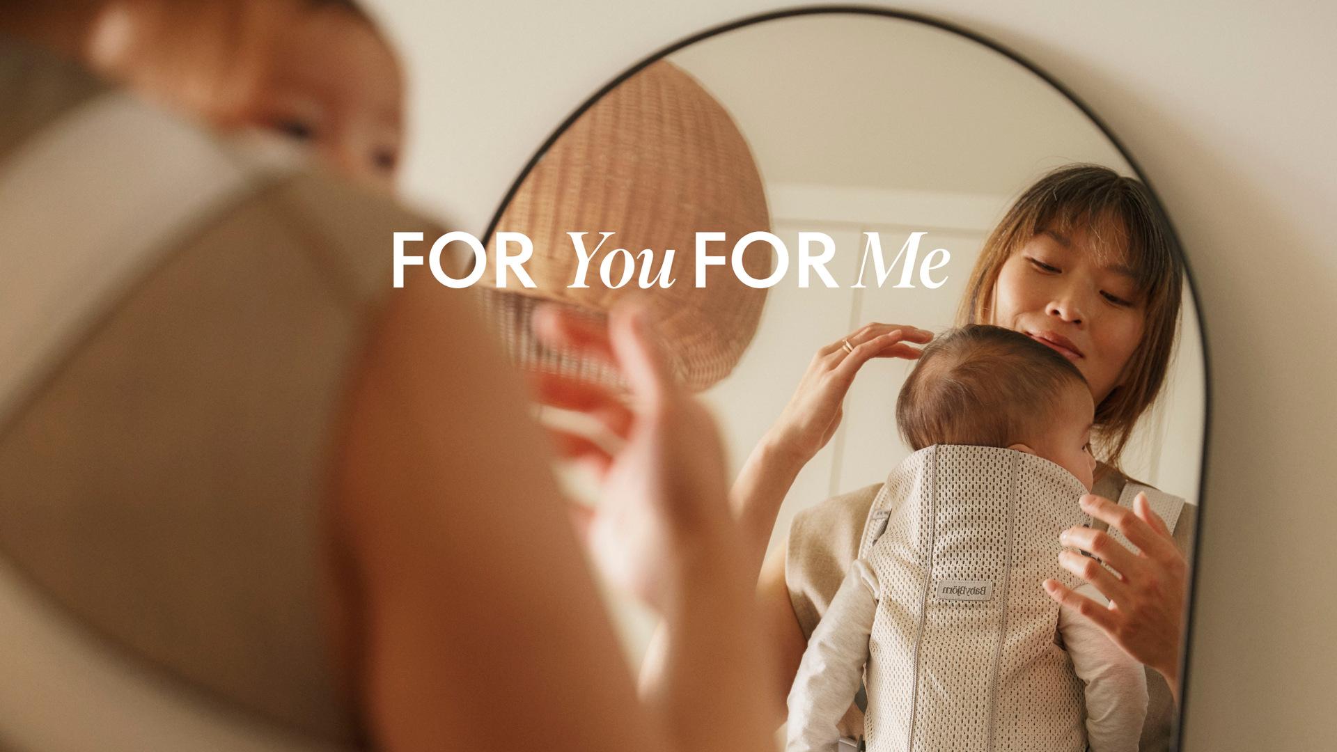 Mother holding a baby in a carrier, looking at their reflection in a mirror with the text "FOR You FOR Me" displayed.
