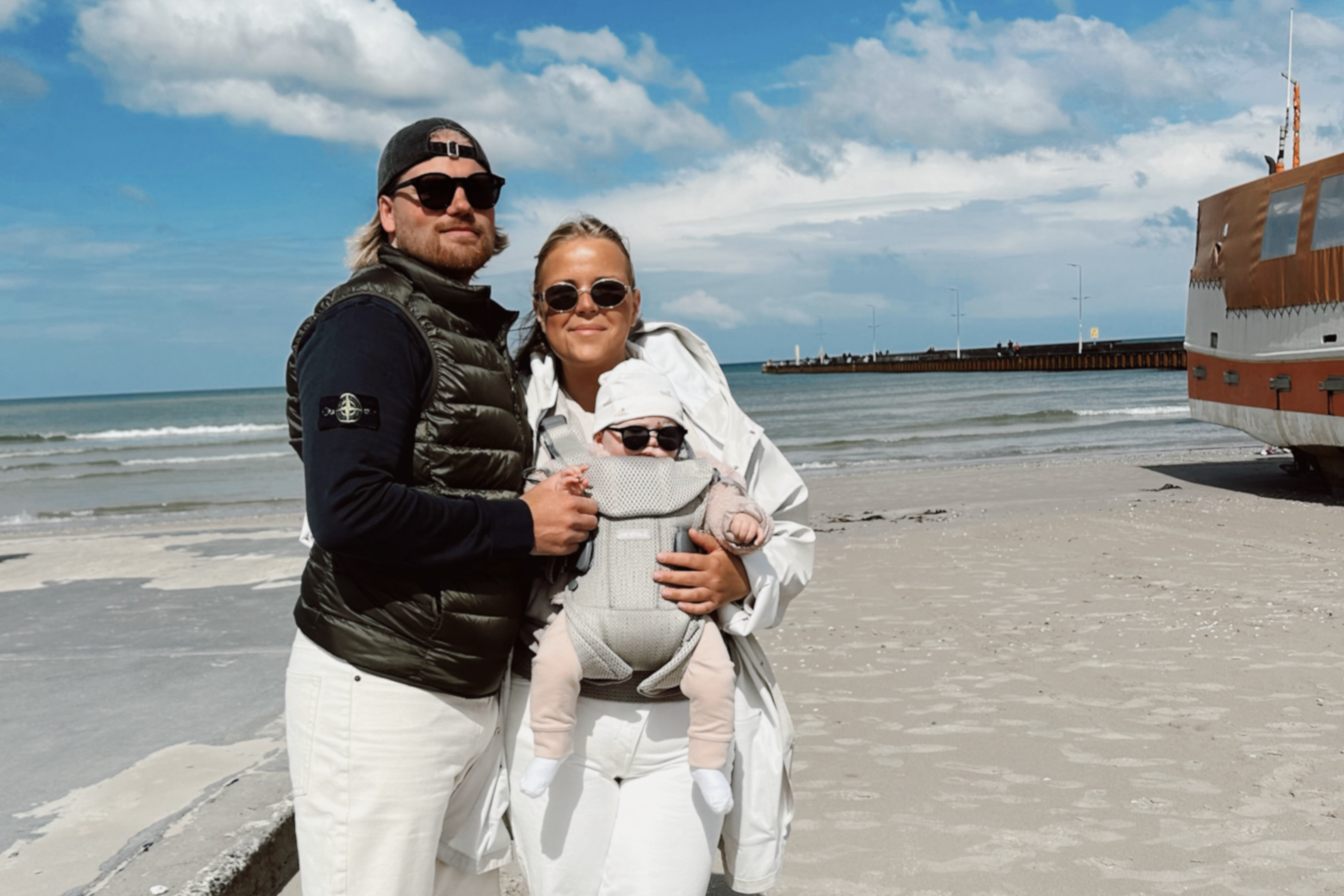 A family standing on a beach; a man and woman wearing sunglasses and casual outfits, holding a baby in a carrier, with the sea and pier in the background.