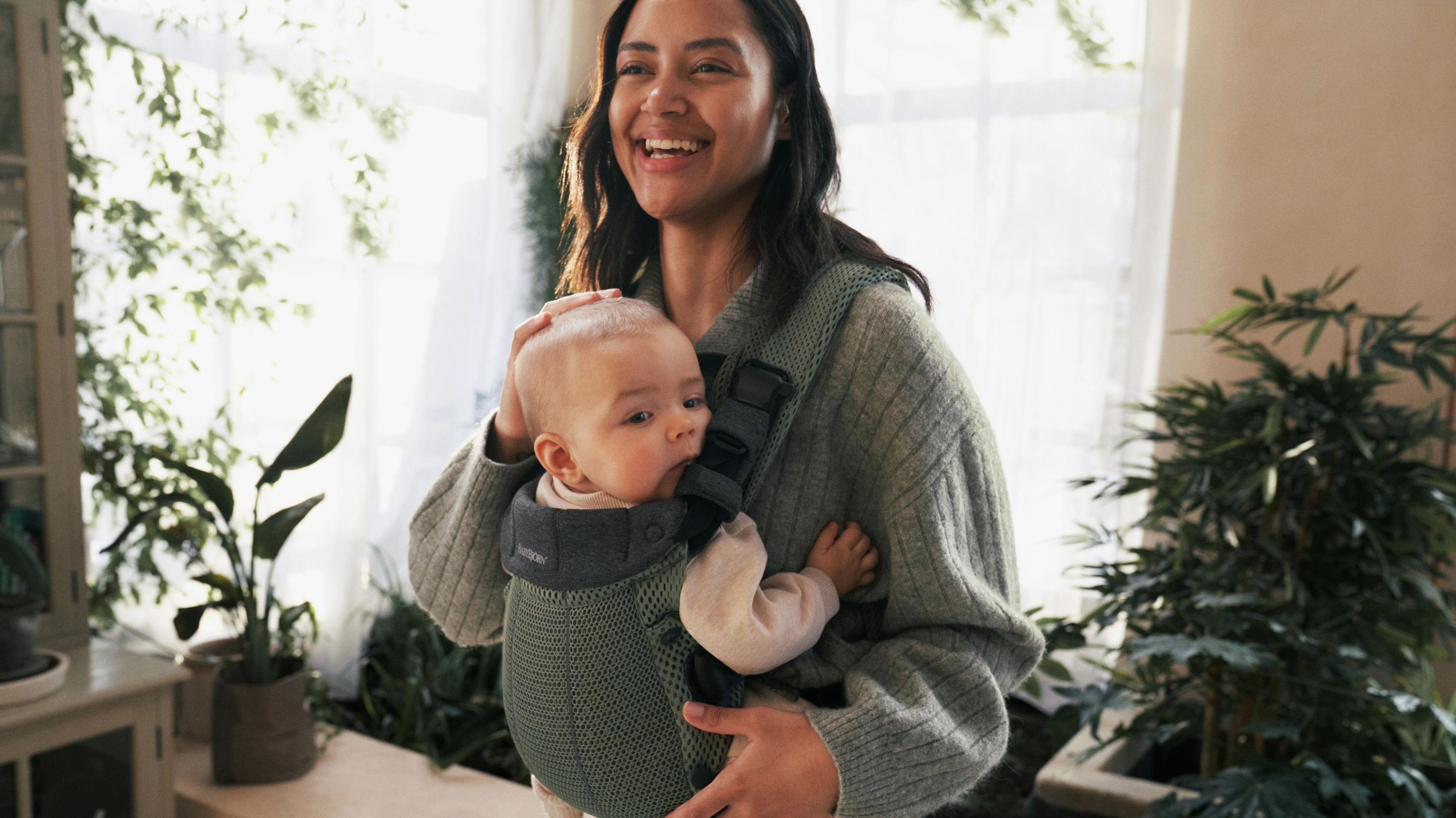 Smiling woman holding a baby in a carrier, surrounded by indoor plants.