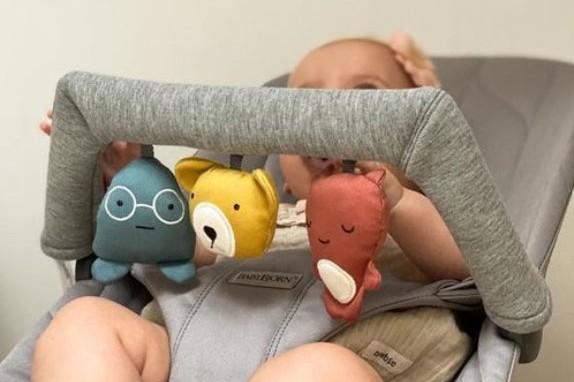 Baby seated in a stroller, holding a bar with three plush toys: a blue figure with glasses, a yellow bear, and a red character.