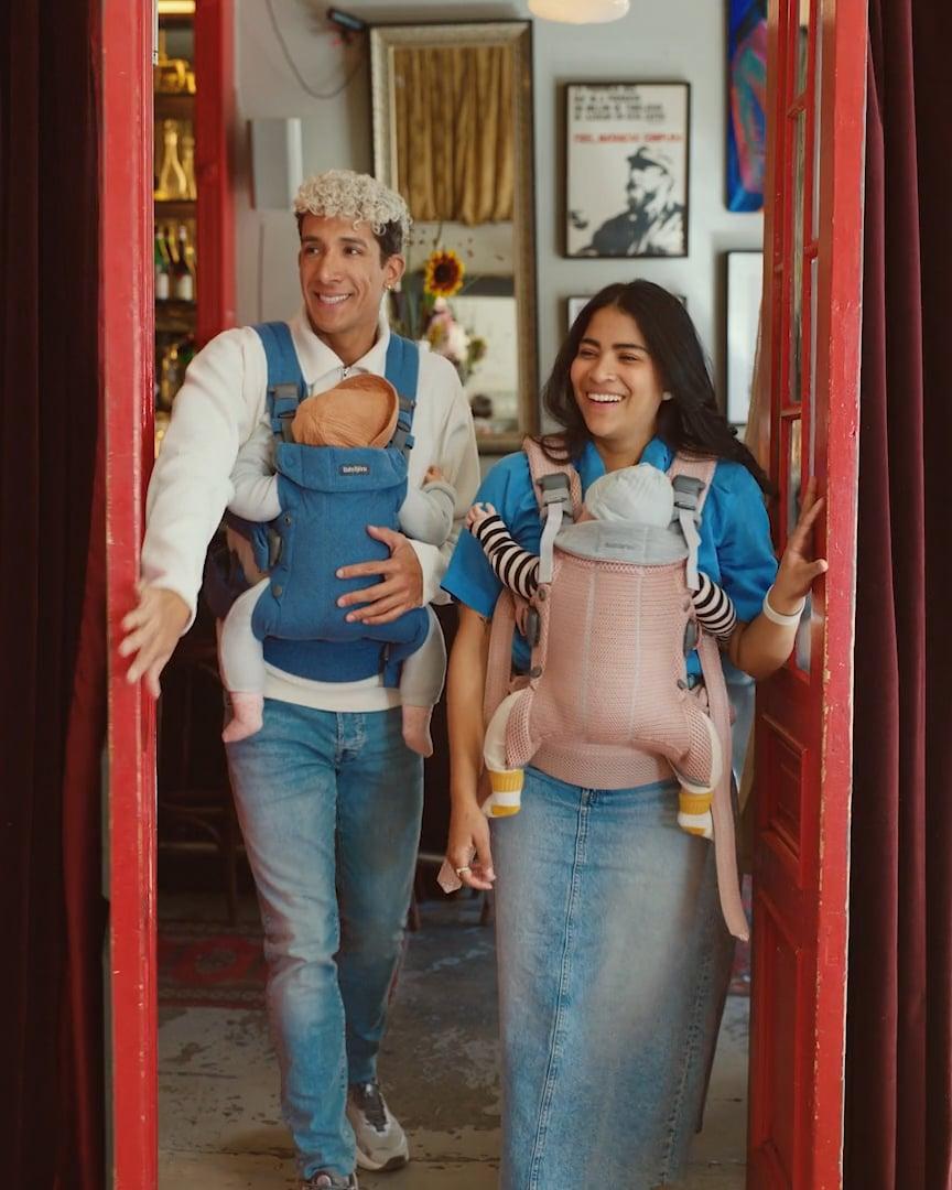 A smiling couple walks through a doorway, each carrying a baby in a front carrier. The man wears a blue carrier, and the woman a pink one.