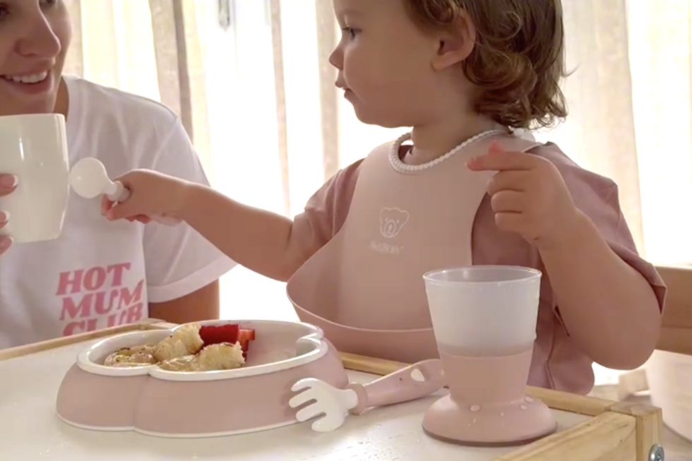 A toddler wearing a bib sits at a table with a pink plate of pancakes and strawberries. An adult next to them holds a cup, smiling.