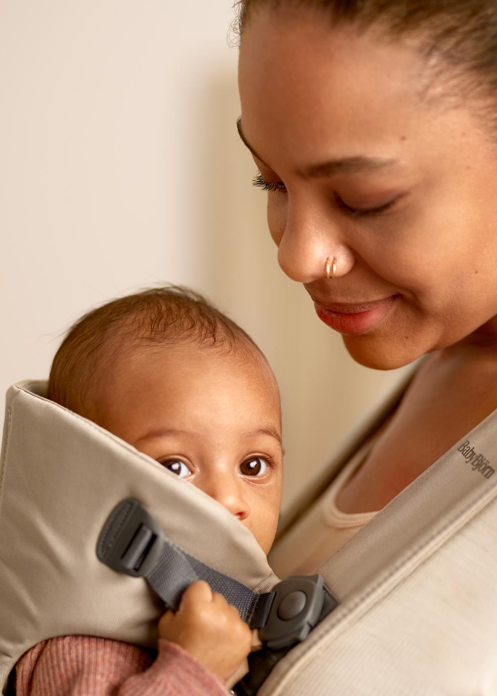 A woman smiles warmly at a baby, who is snuggled in a beige baby carrier on her chest, looking curiously at the camera.