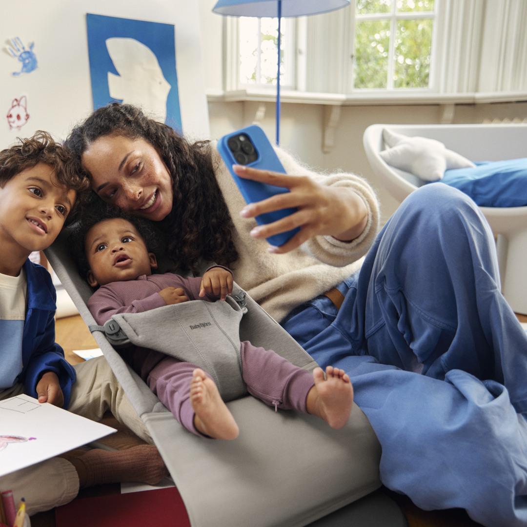 A woman takes a selfie with two children; one is sitting and the other is lying in a baby bouncer. They are in a cozy, well-lit room.