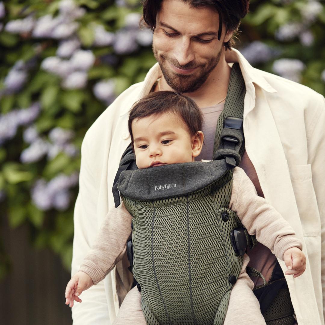 Man smiling at a baby in a green mesh BabyBjörn carrier outdoors, with purple flowers in the background.