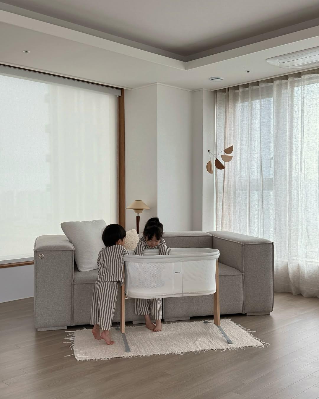 Two children in matching striped pajamas stand beside a small crib in a modern living room with a gray sofa and soft lighting.