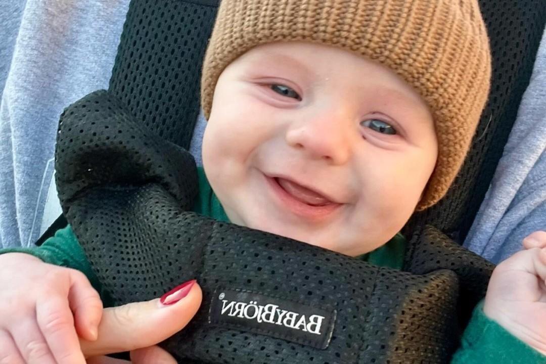 Smiling baby wearing a brown knit hat, sitting in a black BabyBjörn carrier, held by someone with red nail polish.
