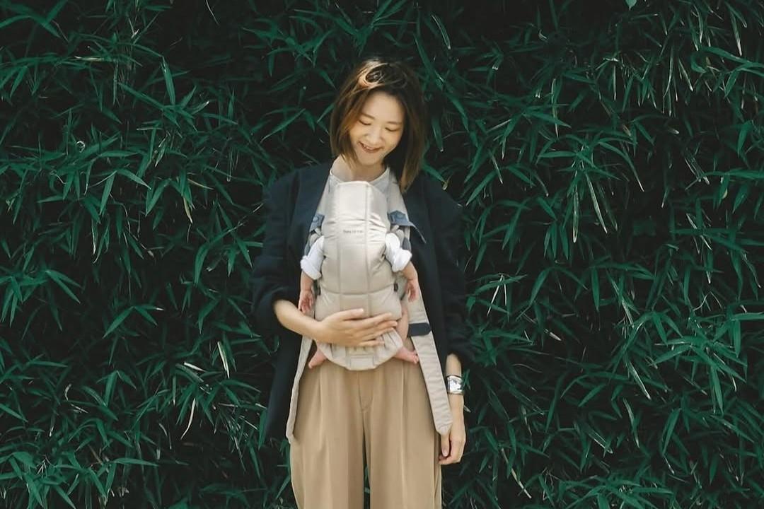 A woman smiles while carrying a baby in a front carrier, standing in front of lush green foliage.