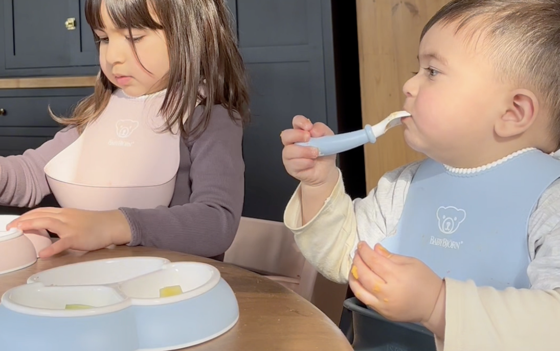 Two young children wearing bibs sit at a table. The older child focuses on food, while the younger one uses a spoon.