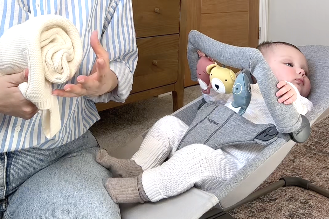 A baby lying in a gray baby bouncer with hanging toys, wearing a white outfit and brown socks, as an adult holds a cream-colored blanket nearby.