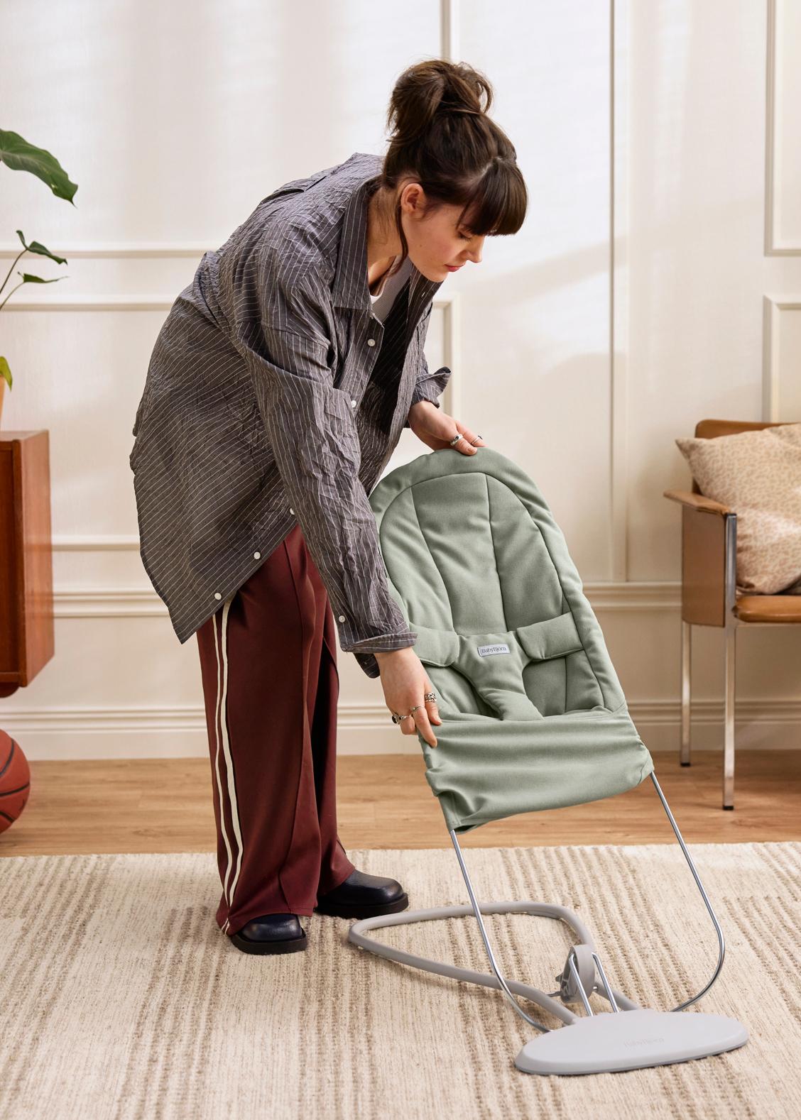 A person adjusting a green baby bouncer in a living room with a basketball in the corner and a chair with cushions.
