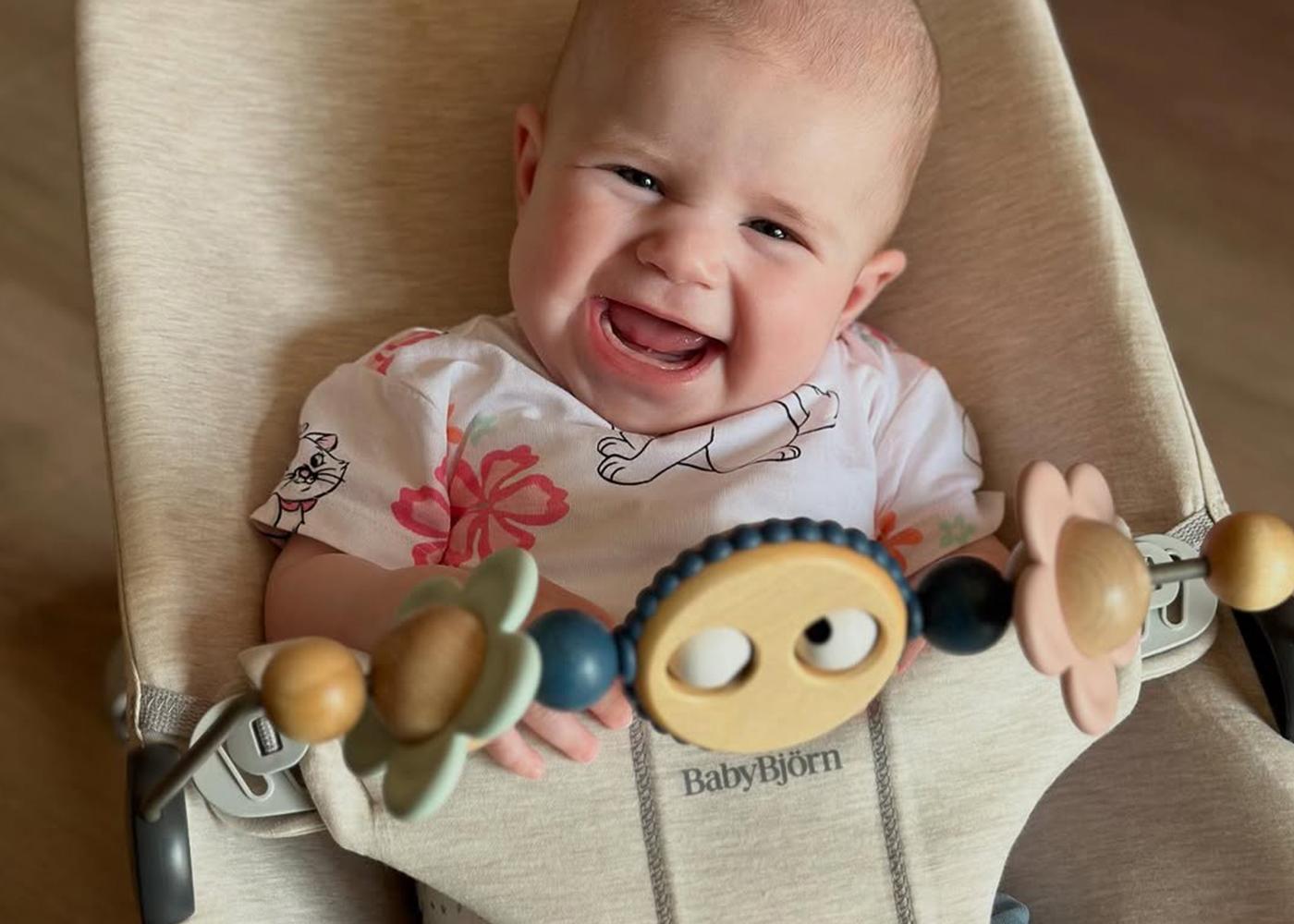 A smiling baby in a floral shirt sits in a beige BabyBjörn bouncer, holding a toy with colorful flower shapes.