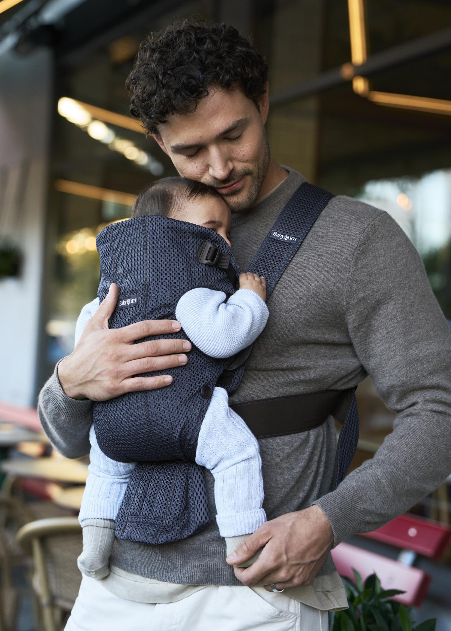 A man gently holds a baby in a navy blue carrier, standing outdoors with blurred background of a cafe, showing a tender moment.
