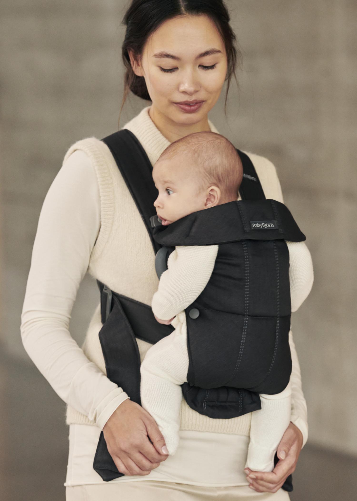 A woman wearing a beige sweater holds a baby in a black front-facing carrier. The baby is gazing to the side while the woman looks down.