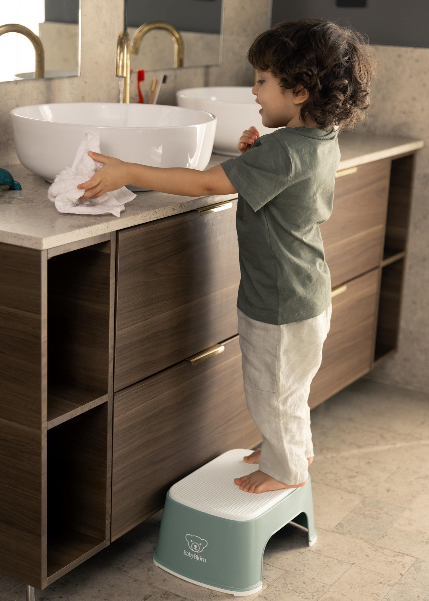 A child stands on a stool, brushing teeth at a bathroom sink with a towel in hand, wearing a green shirt and white pants.