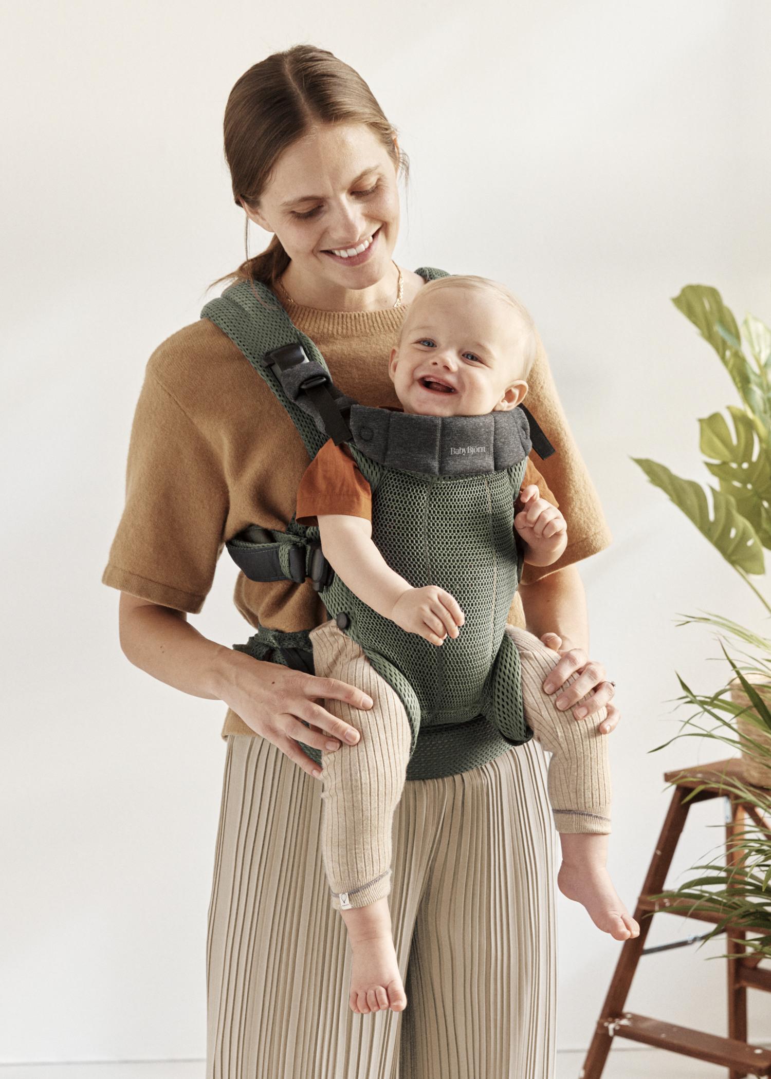A smiling woman in a brown top carries a happy baby in a green mesh baby carrier. The setting is bright with a plant in the background.