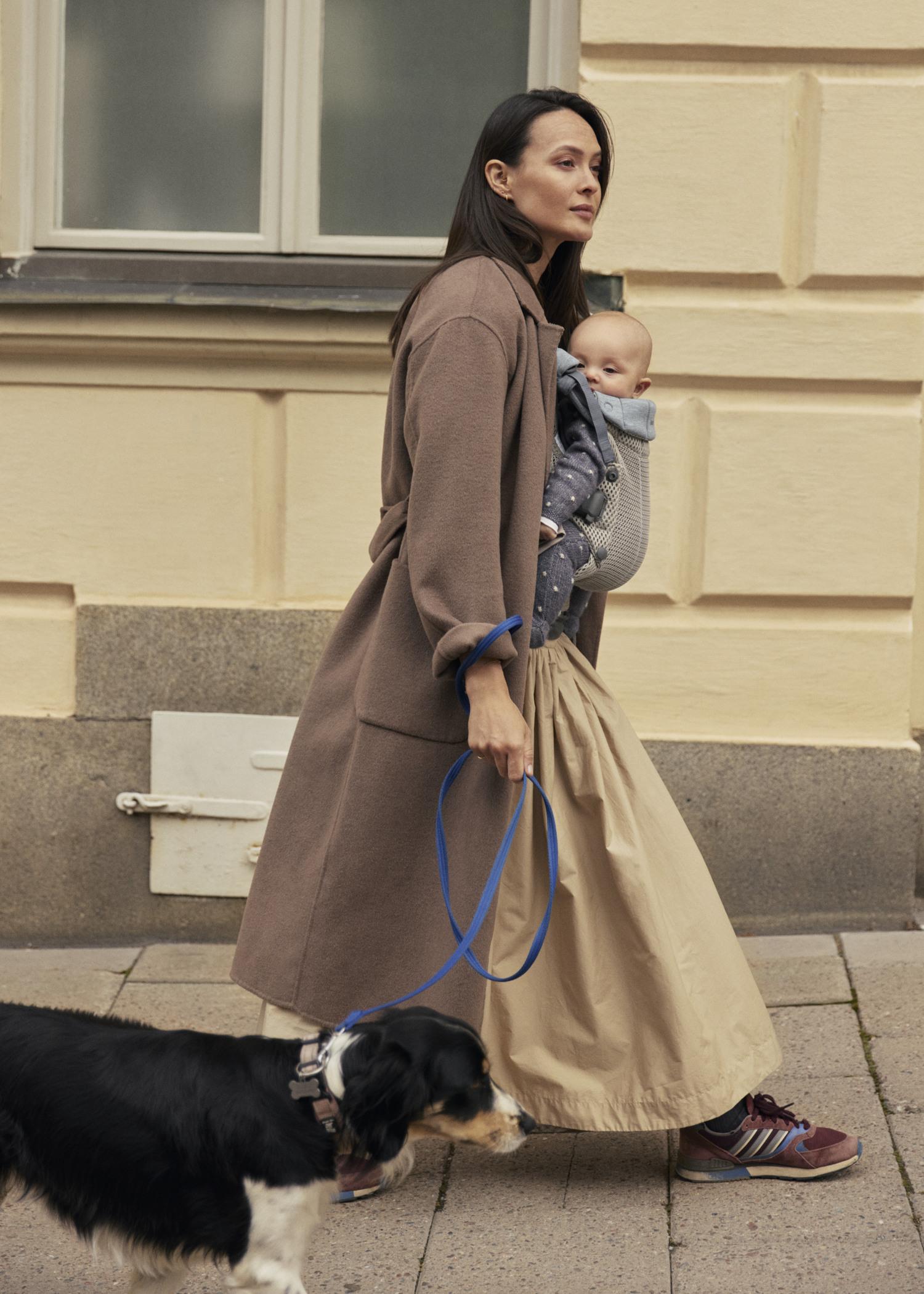 A woman walks outside, carrying a baby in a front baby carrier and holding a dog's leash. She wears a long coat and skirt. Scandinavian style.