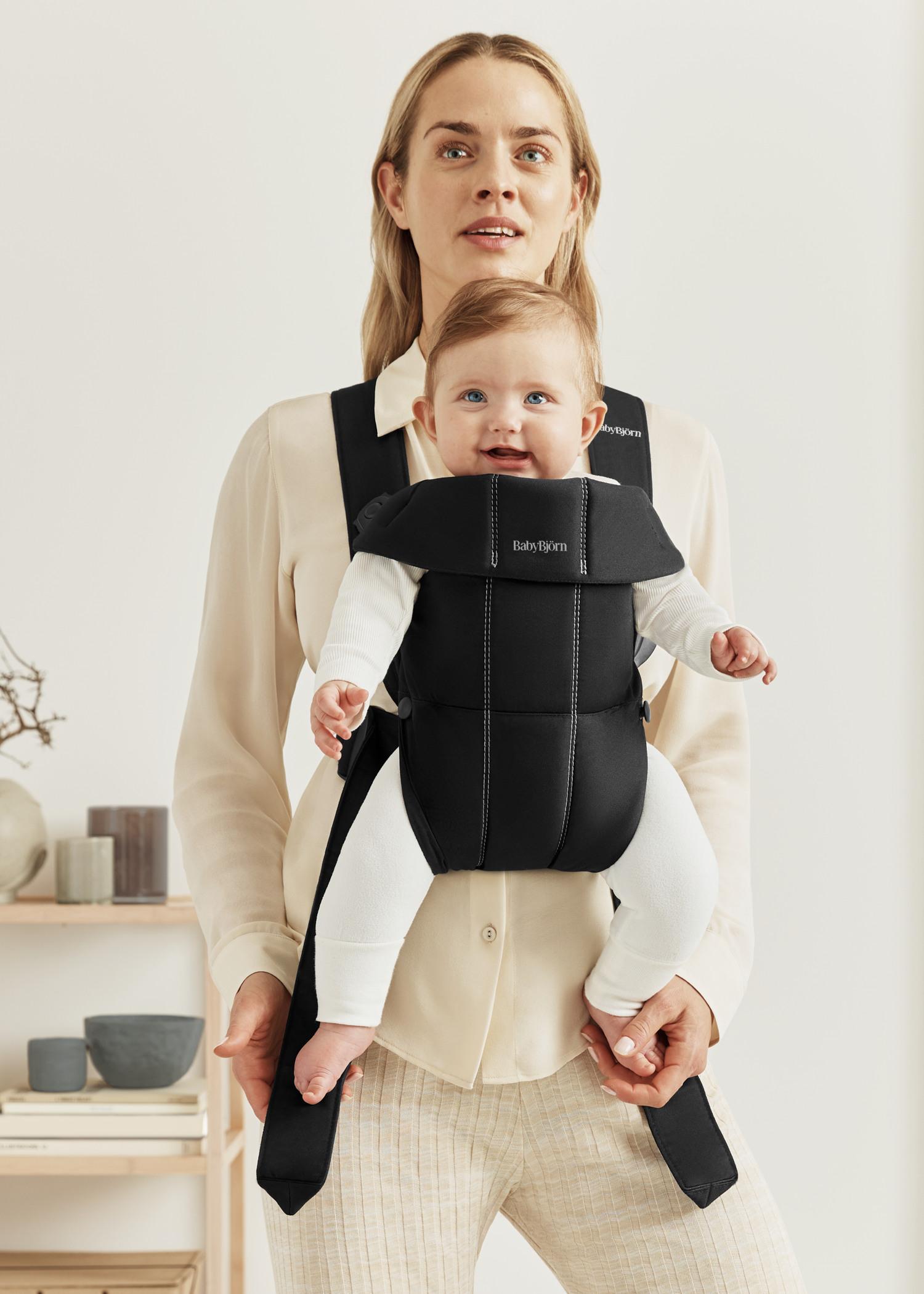 Adult holding a smiling baby in a black carrier, standing indoors with light background and minimalist decor.