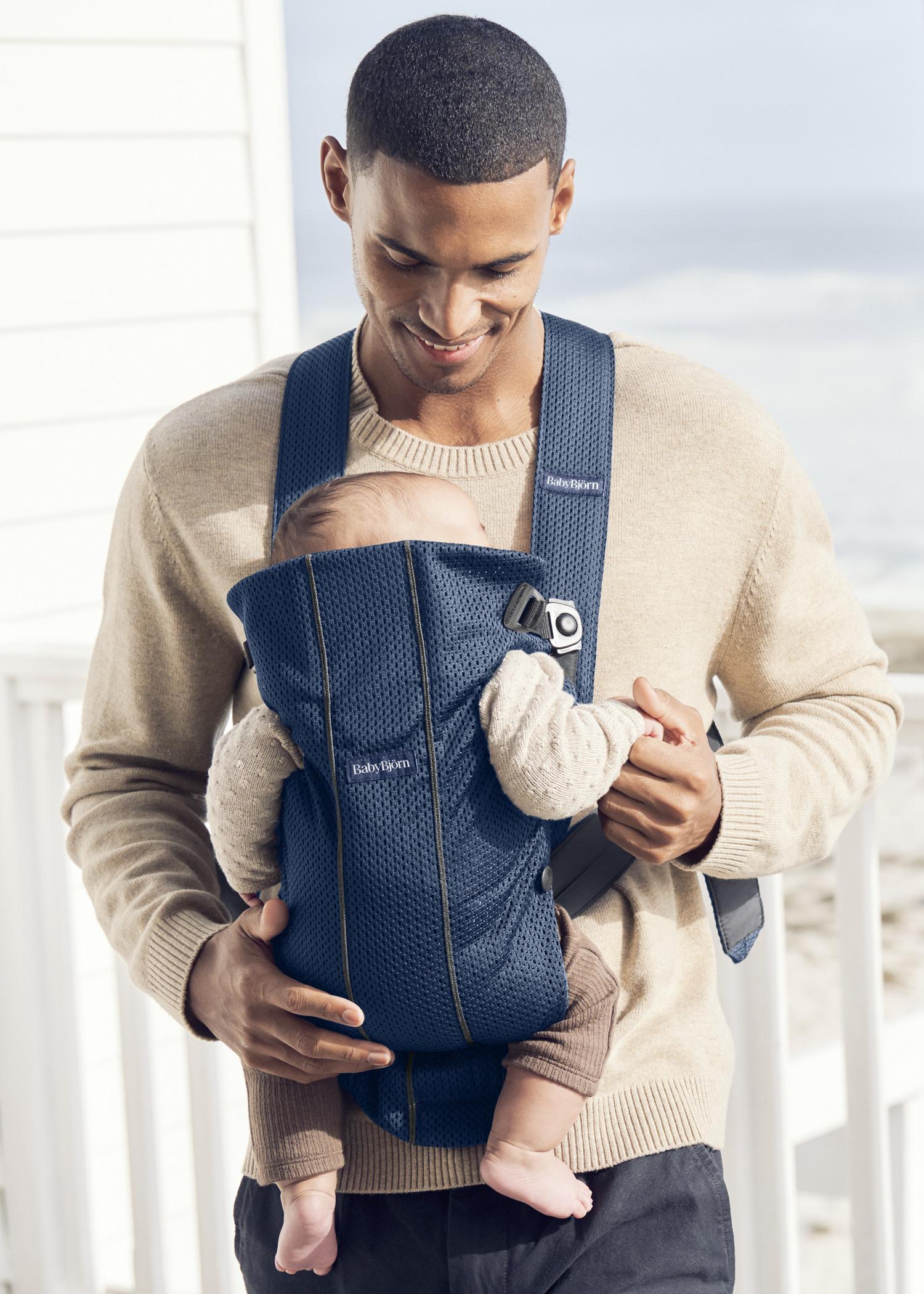 A person smiles while carrying a baby in a blue baby carrier. They are standing outside with a blurred background of a beach and building.