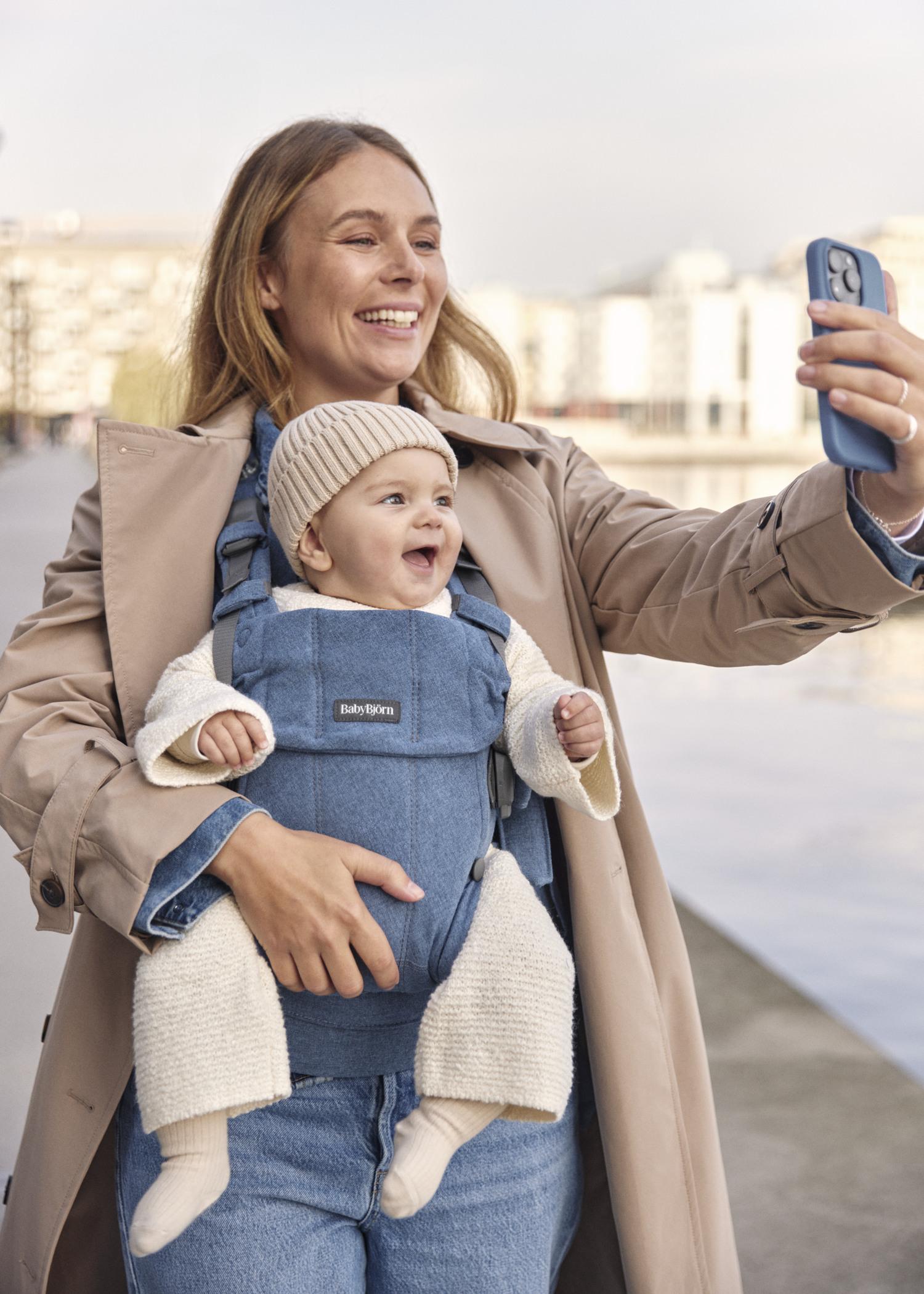 Smiling woman in a trench coat takes a selfie with a happy baby in a blue carrier, near a waterfront.