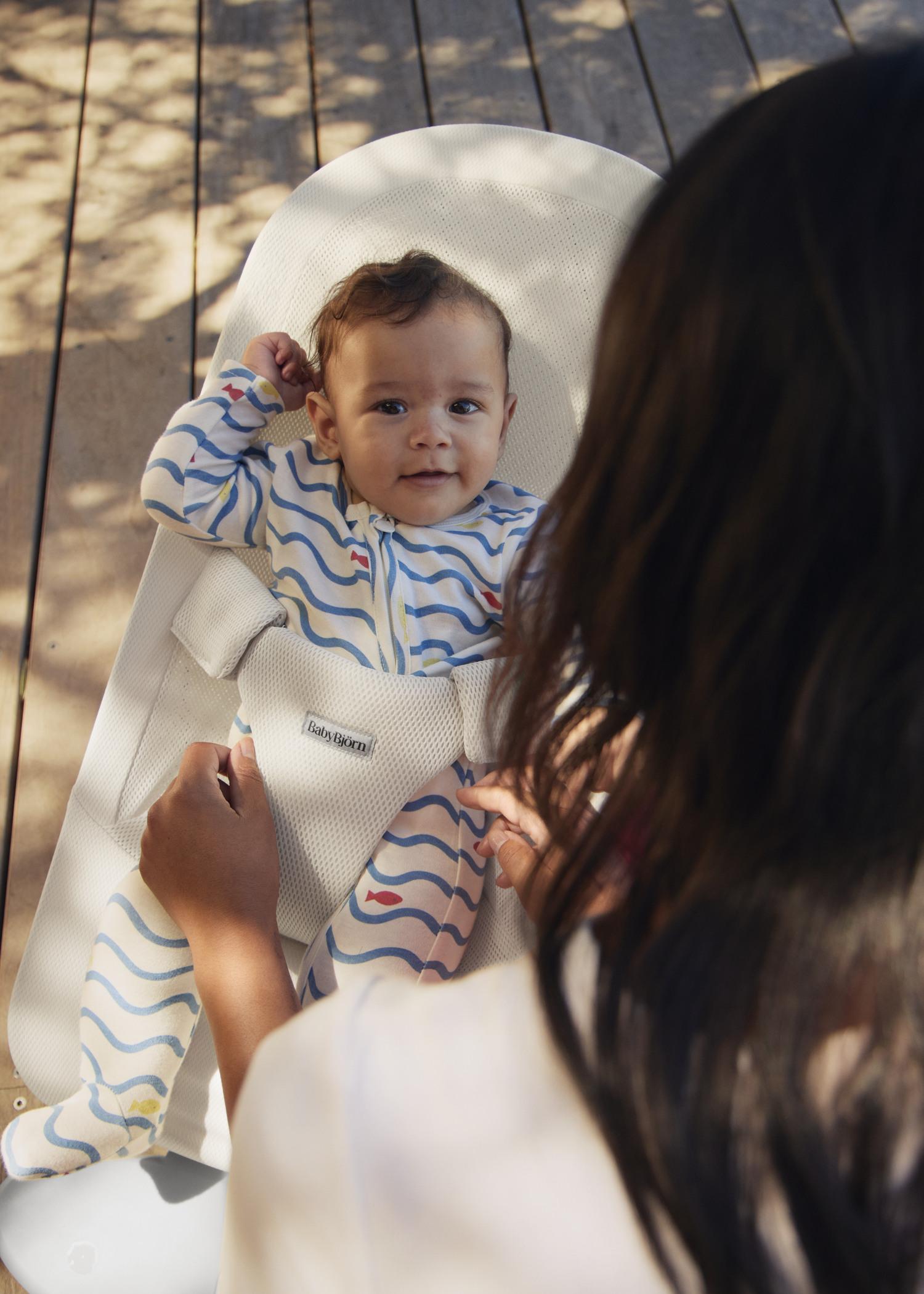Baby in a patterned onesie lying in a bouncer chair, smiling up at an adult with hands gently holding the bouncer's straps.