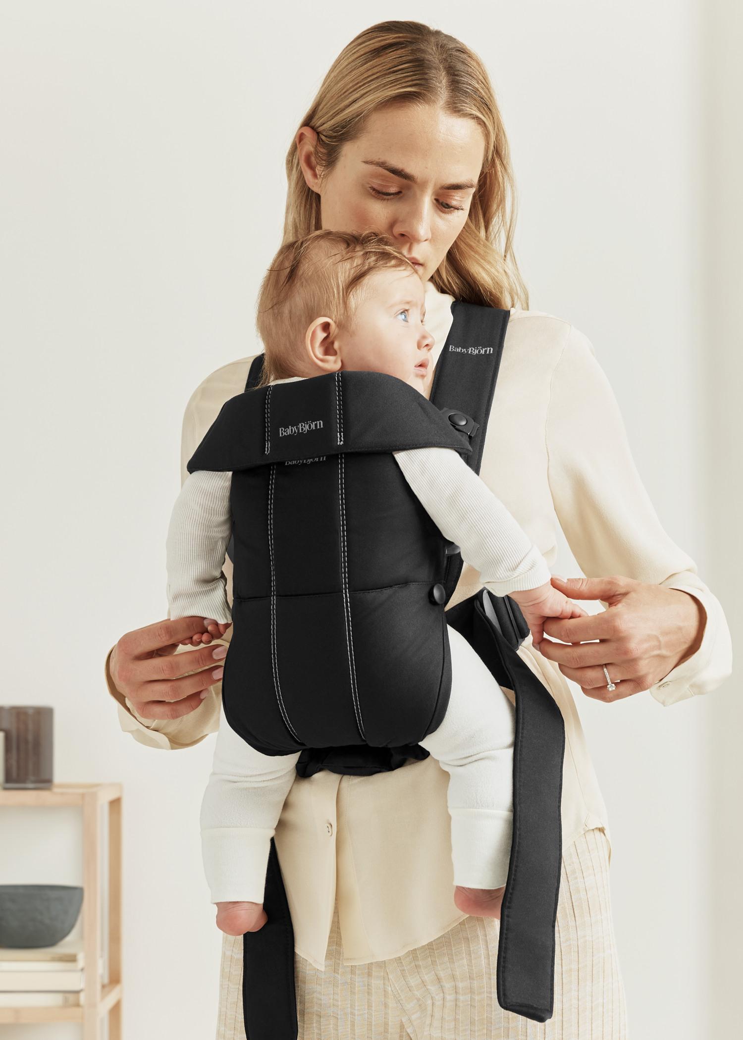 A woman holding a baby in a black carrier, both dressed in light, neutral clothing, standing in a bright, minimalistic room.