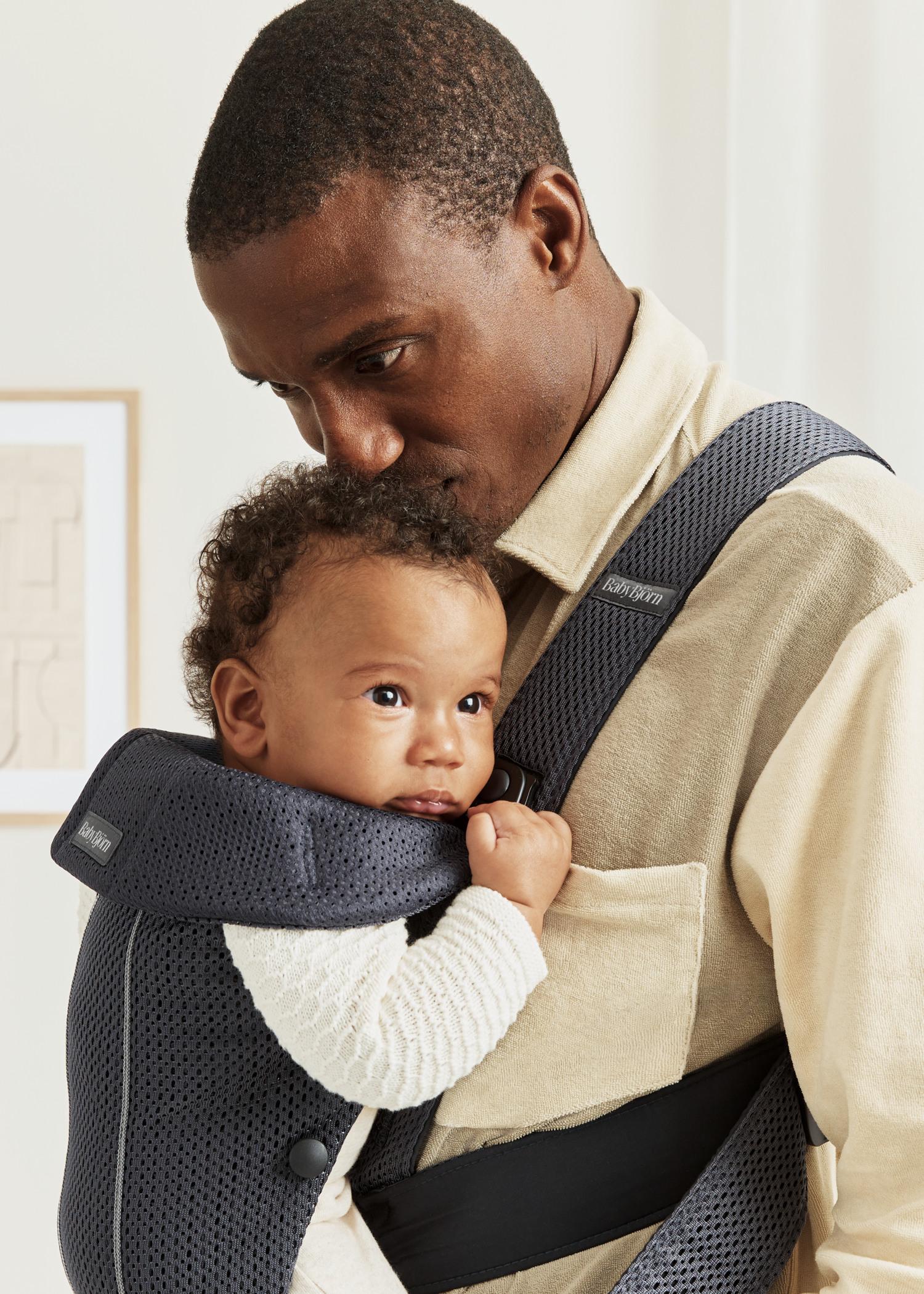 A man lovingly looks at a baby in a black carrier. The baby is held close to his chest, wearing a white sweater in a cozy, light-filled room.