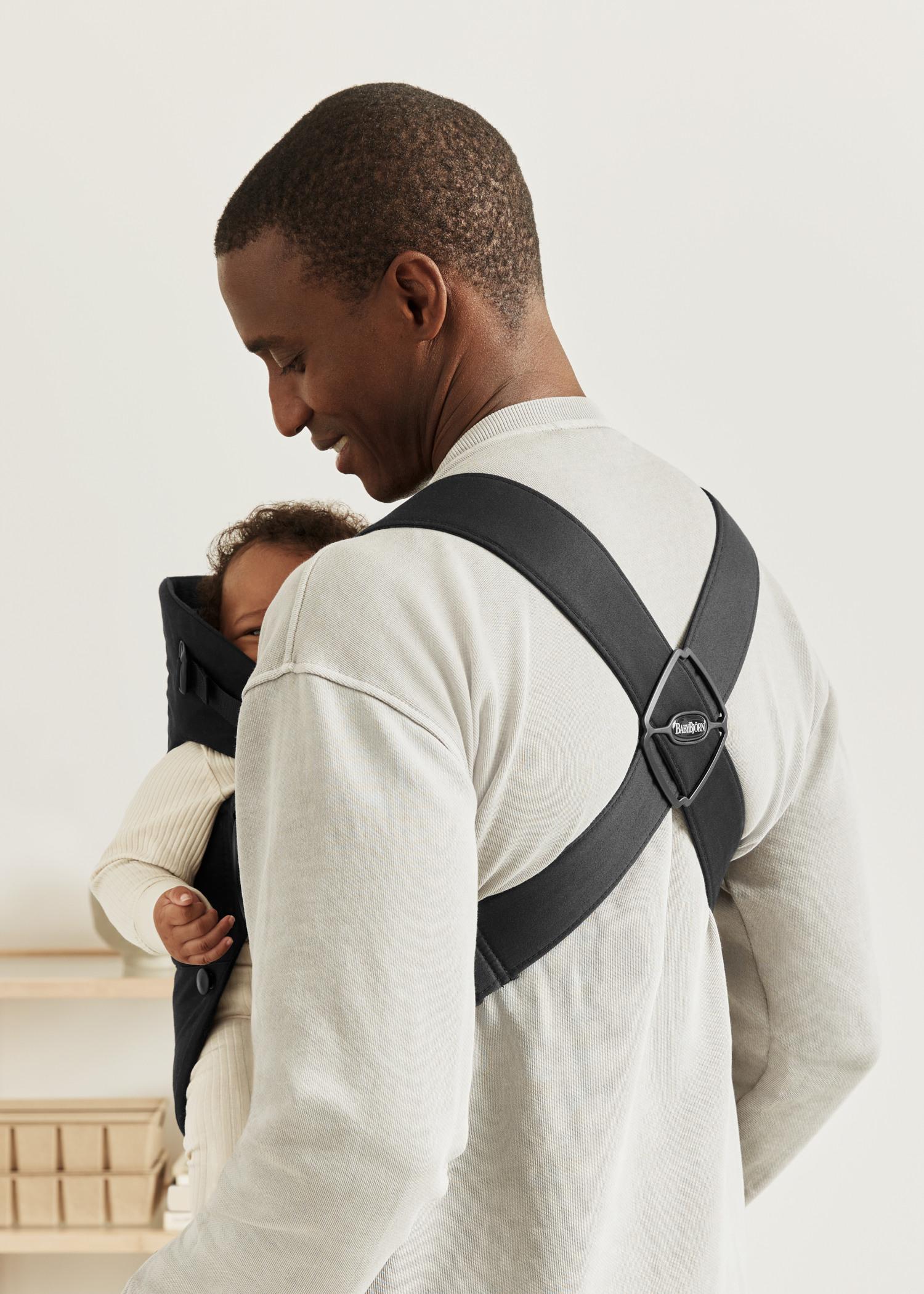 A man wearing a light-colored shirt uses a black baby carrier to hold an infant. The baby rests comfortably against his chest.