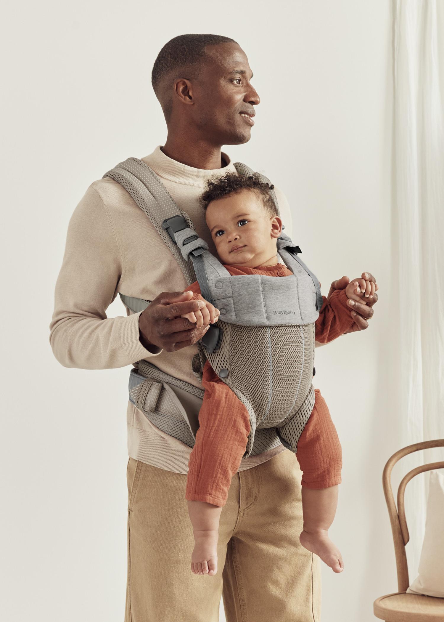 A man smiles while holding a baby in a gray beige carrier. The baby, wearing a rust-colored outfit, looks comfy.