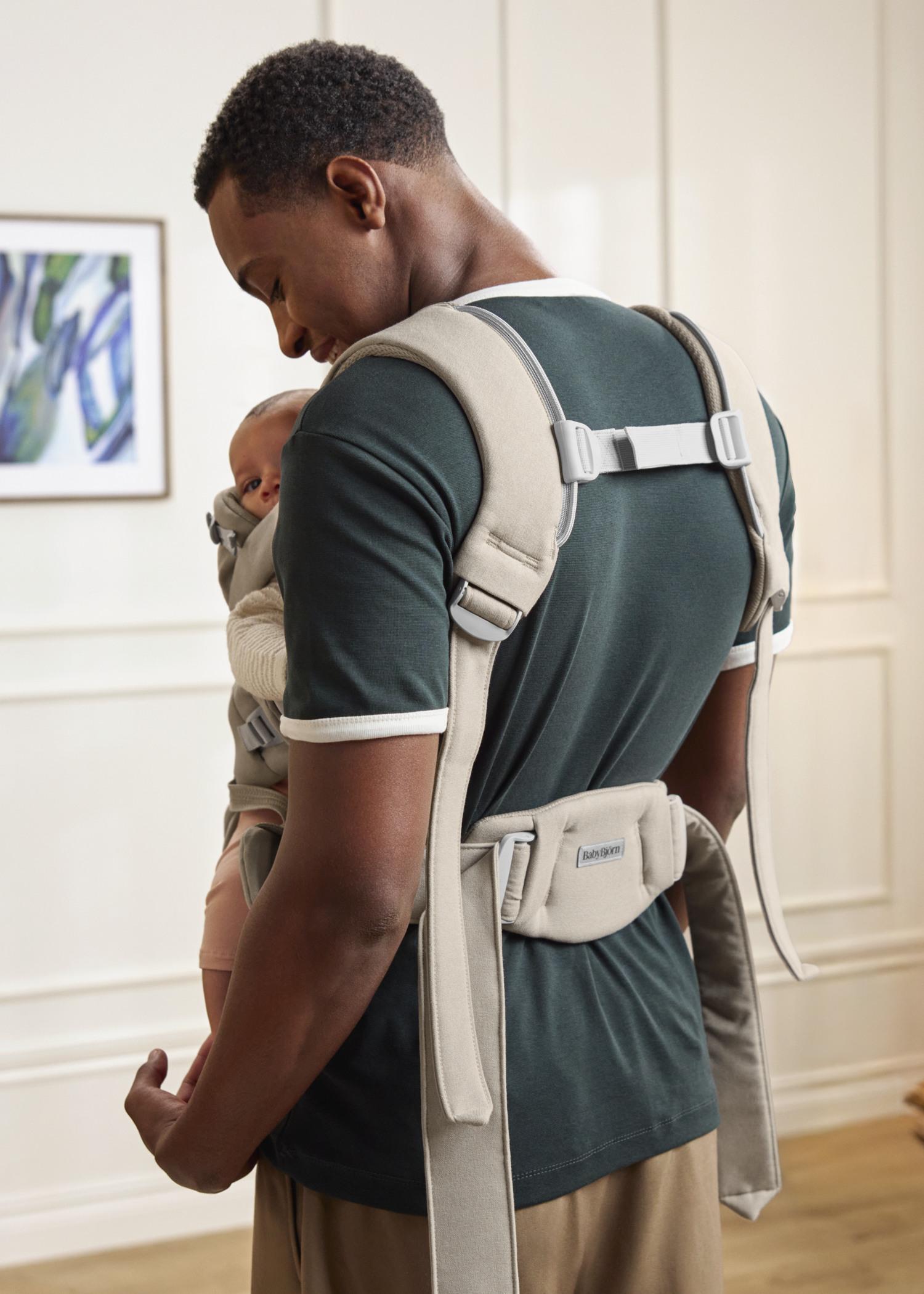 Man wearing a green shirt and khaki pants carries a baby in a beige baby carrier in a bright room with a painting on the wall.