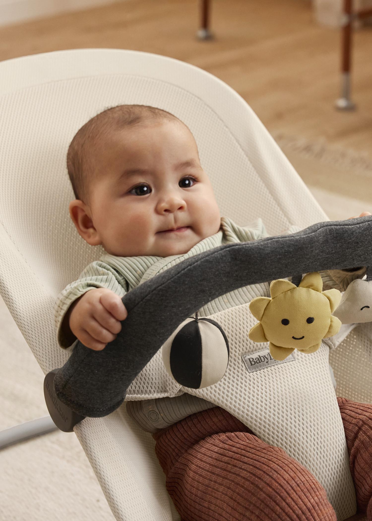 A baby sitting in a white fabric bouncer, wearing a green shirt and brown pants, holding a toy bar with a smiling sun plush toy attached.