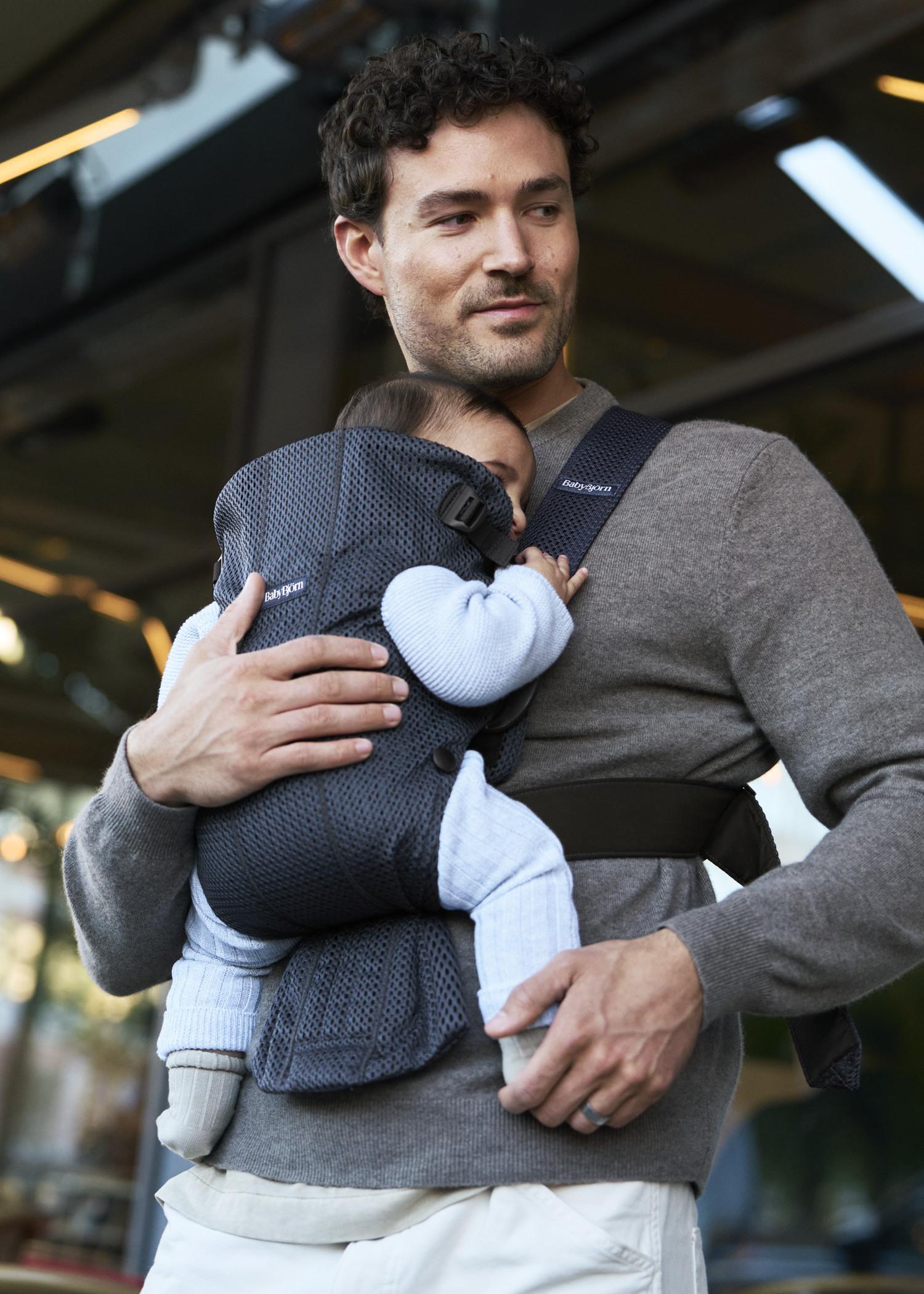 A man wearing a gray sweater carries a sleeping baby in a blue baby carrier while standing outside.