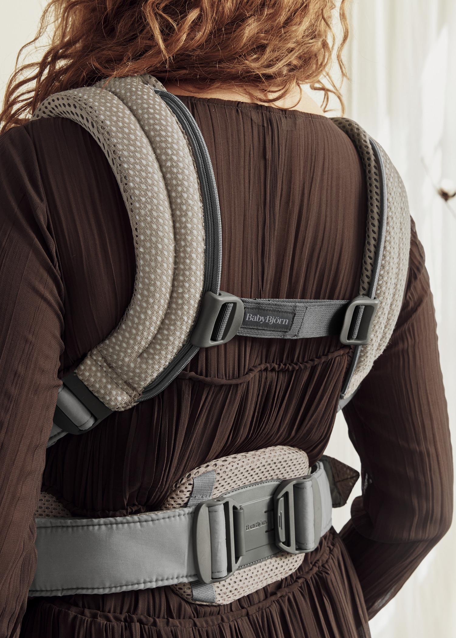 Person wearing a BabyBjörn baby carrier, viewed from the back, with padded straps for back support and a dark brown outfit.