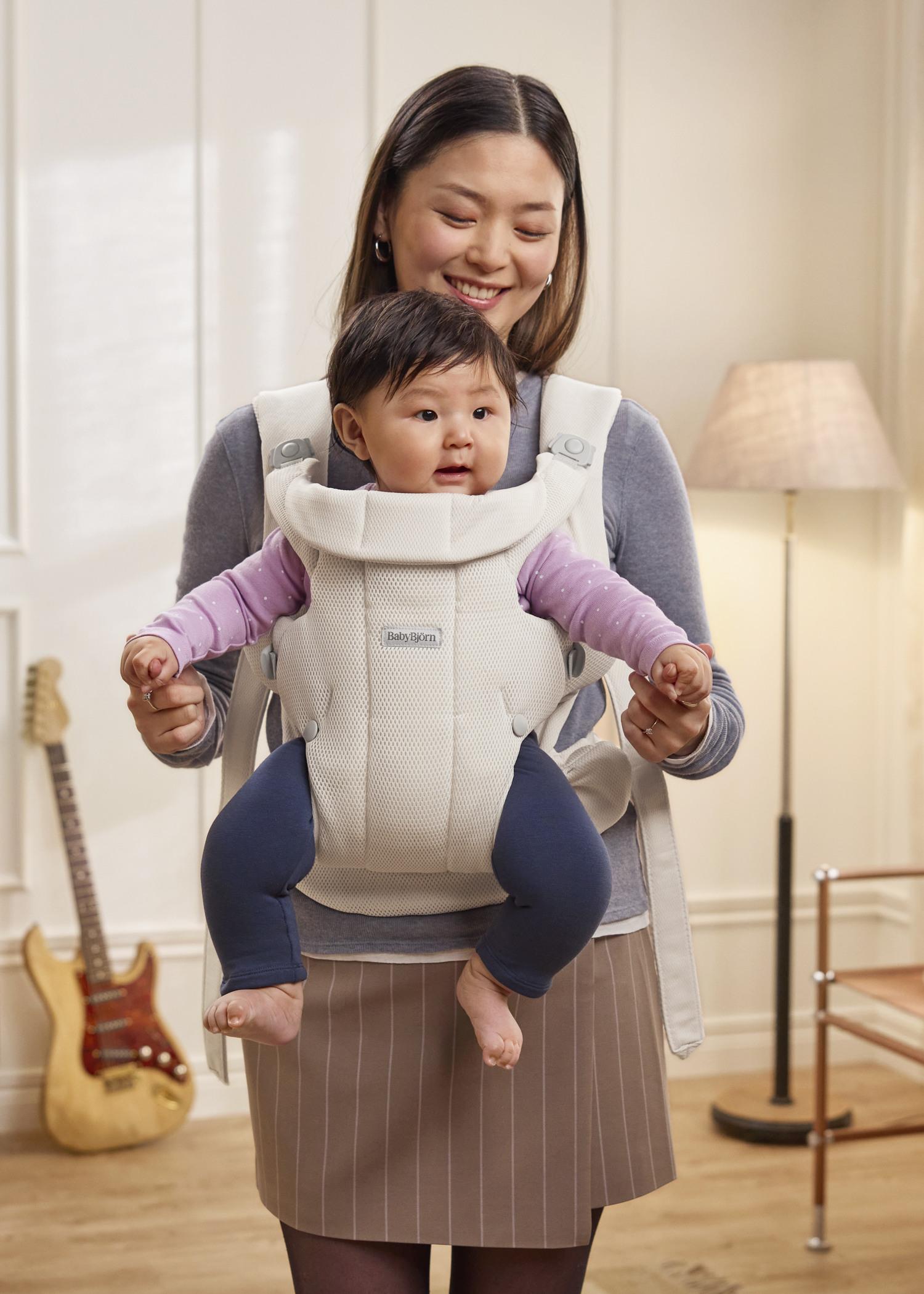 A woman smiling at a baby in a baby carrier. The baby is wearing a pink top and blue pants. A guitar and lamp are in the background.
