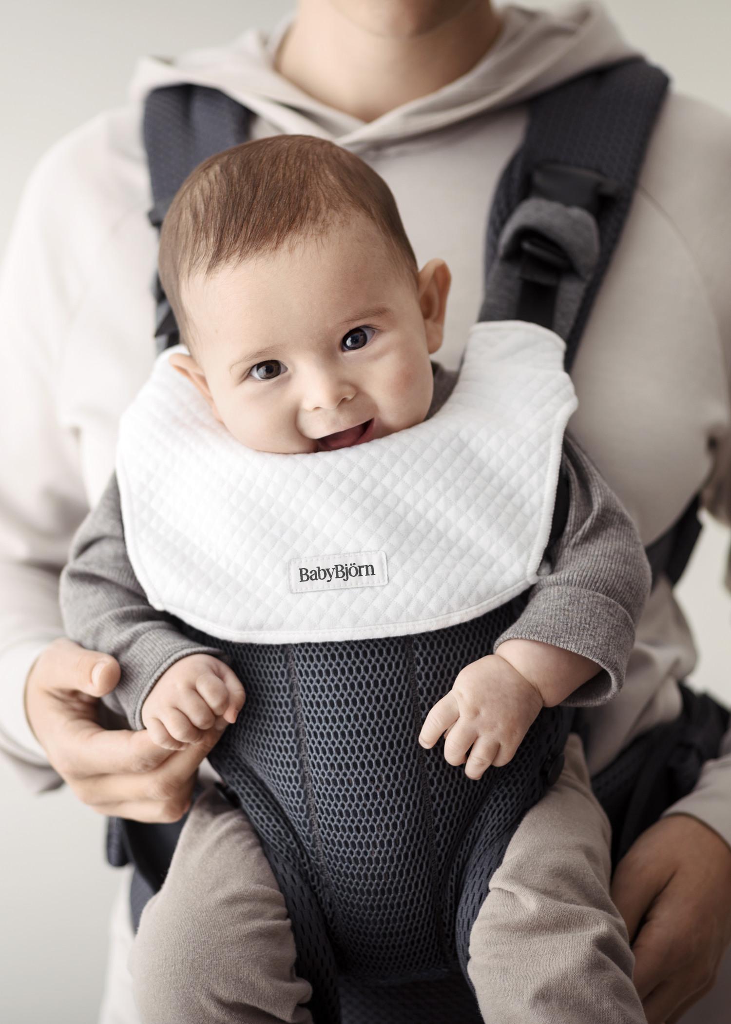 Smiling baby in a gray baby carrier, wearing a white bib, held by an adult in a beige hoodie.