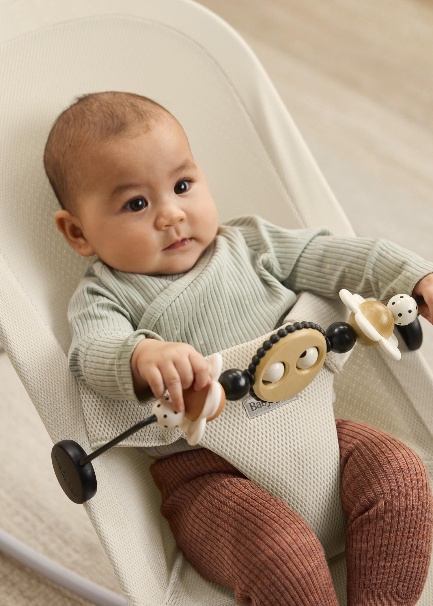 A baby in a light green outfit sits in a bouncer chair, holding a toy bar with various shapes and colors, looking intently forward.