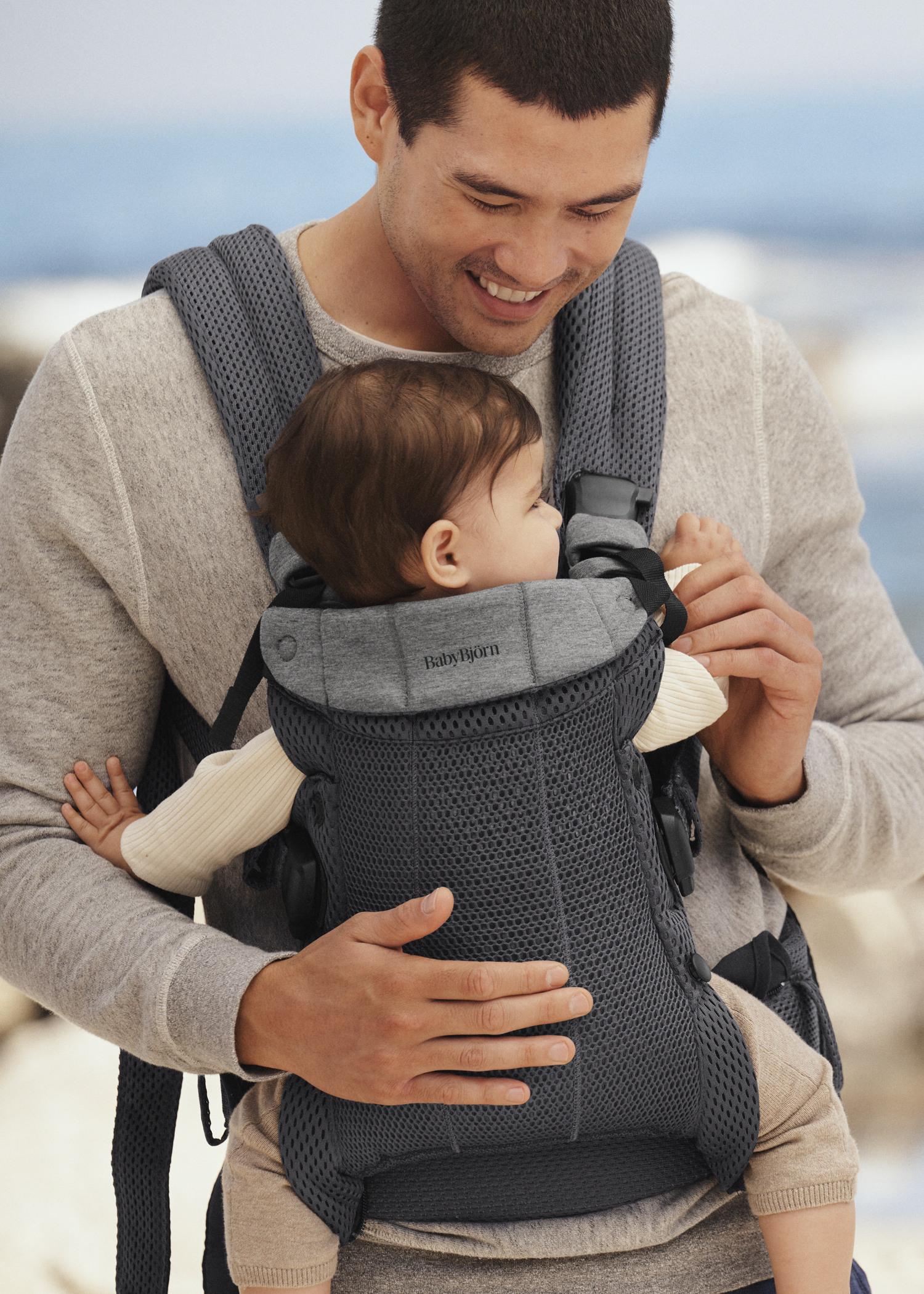A smiling man holds a baby in a gray baby carrier, both dressed in light-colored clothing, with a blurred outdoor background.