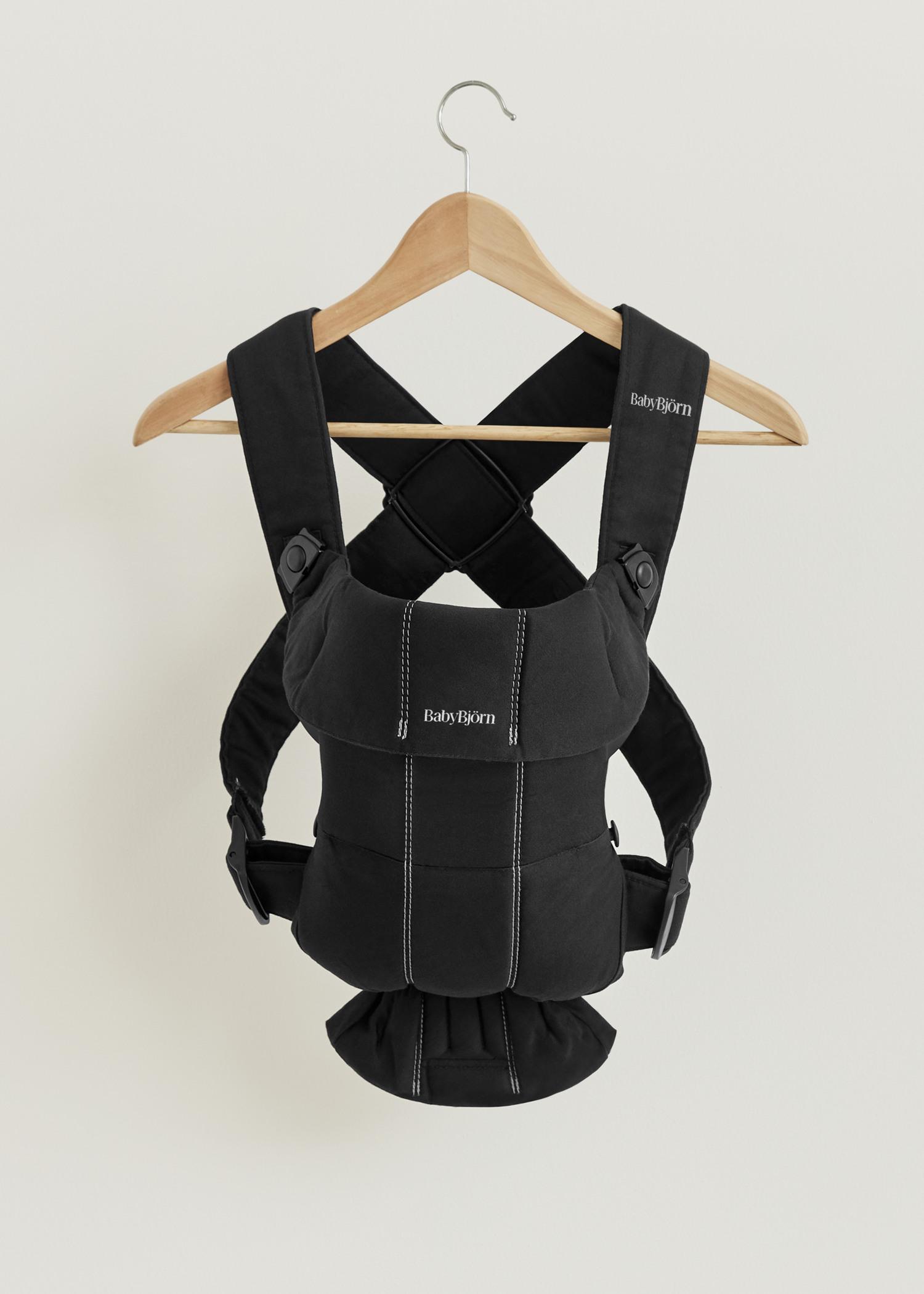 Black ergonomic baby carrier with adjustable straps hanging on a wooden hanger against a white background.