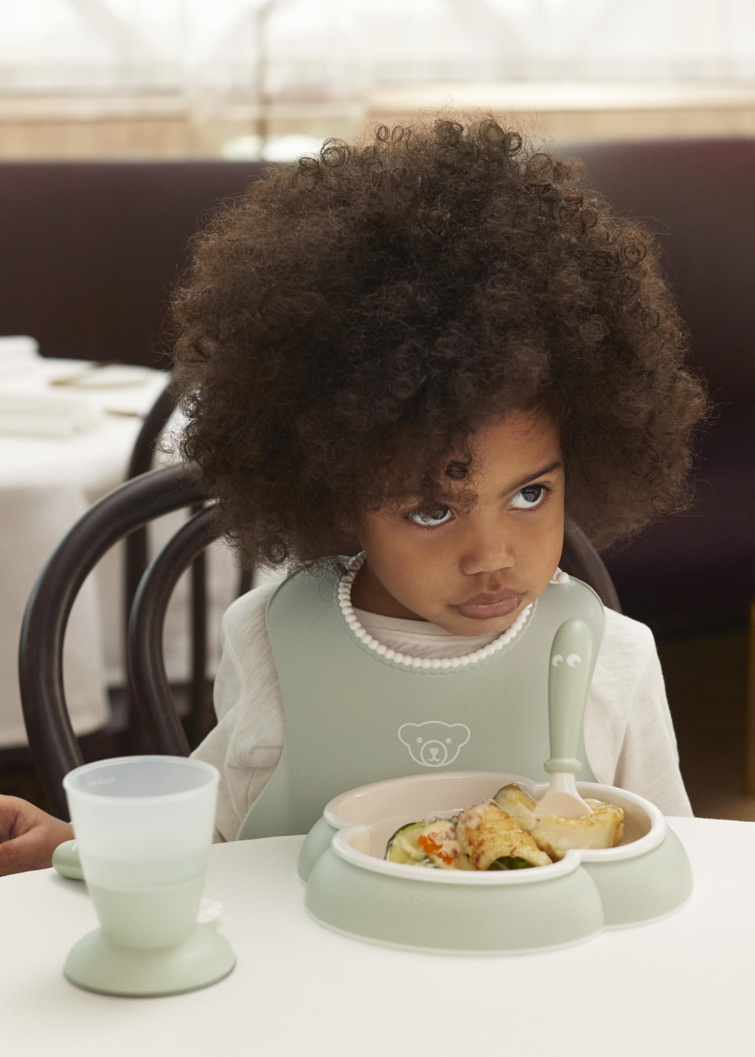 A child with curly hair wearing a bib sits at a table with a plate of food and a sippy cup in front of them, looking thoughtfully to the side.