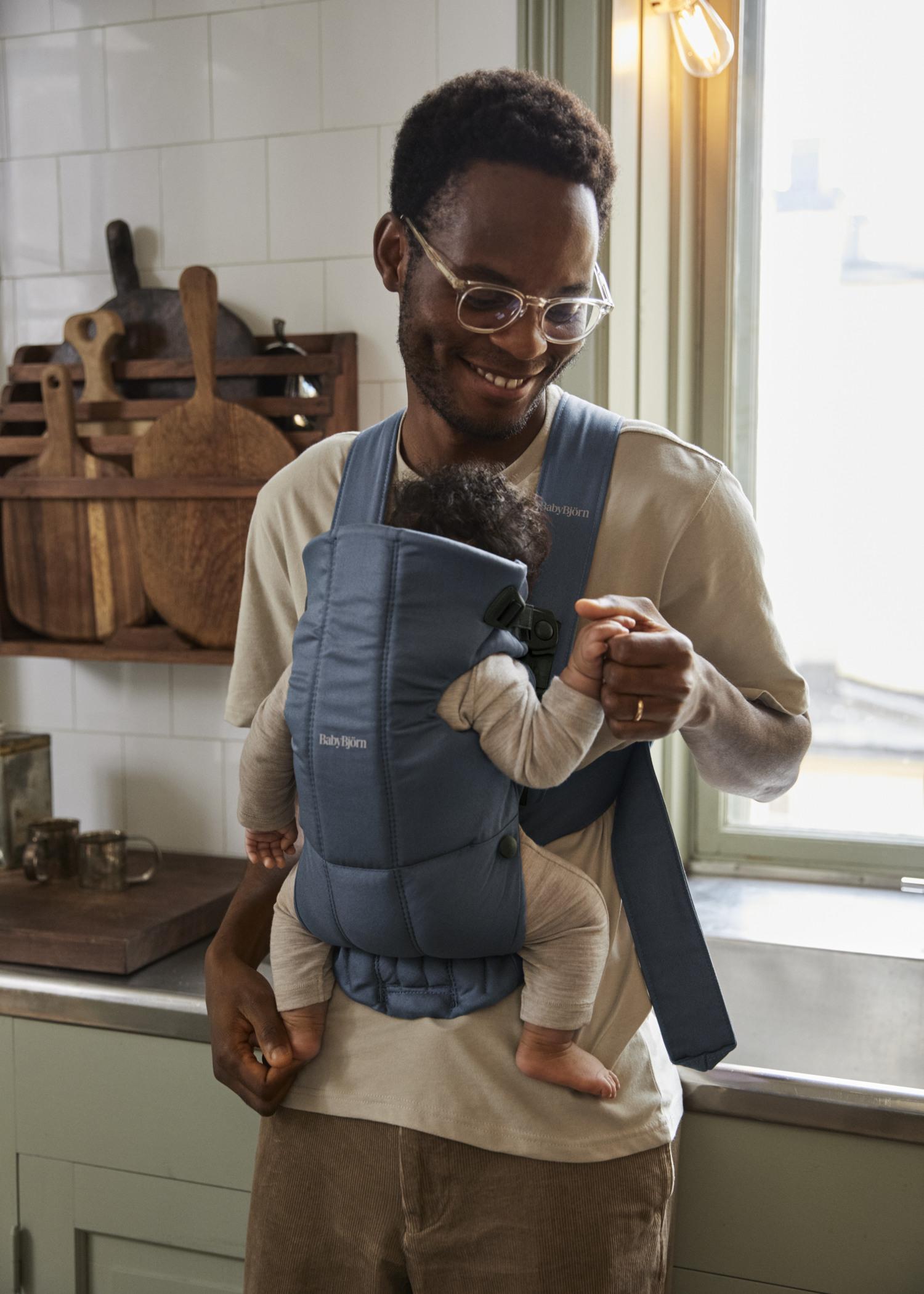 A man wearing glasses smiles while holding hands with a baby in a blue baby carrier inside a kitchen.